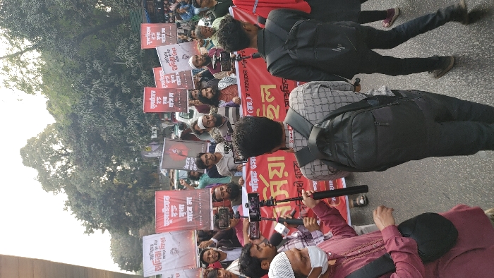 This daytime image depicts a street protest or demonstration in Dhaka, Bangladesh. A dense crowd of predominantly adult males is gathered, many holding red and white banners with Bengali text. One prominent red banner stretches across the middle ground, displaying white Bengali text, including the phrases "শারীরিক ও মানসিক" (physical and mental) and "ইল" (possibly part of a larger word). Other banners held by individuals in the background feature Bengali text such as "শাদিয়া" (Shadia/Sadia), "ইন্ডিয়া" (India), and phrases like "অপমান বরদাস্ত করা হবে না" (insult will not be tolerated), along with some partial English text like "Insult is not ever."
Several participants are actively documenting the event; multiple individuals hold smartphones mounted on monopods or selfie sticks, pointing towards the center of the gathering. One man in the left foreground wears a maroon shirt, a white prayer cap (topi), and a white face mask, holding a black microphone towards the crowd. Another individual in the immediate foreground, with his back to the camera, wears a plaid shirt and a black backpack. To his right, another person, also with their back to the camera, wears a black top and a black backpack.
The environment shows a paved road surface with a significant amount of green foliage and trees in the background, suggesting an urban street setting. A light-colored overhead structure, possibly an elevated road or flyover, is visible on the far left. Distant vehicles are faintly visible behind the protestors.