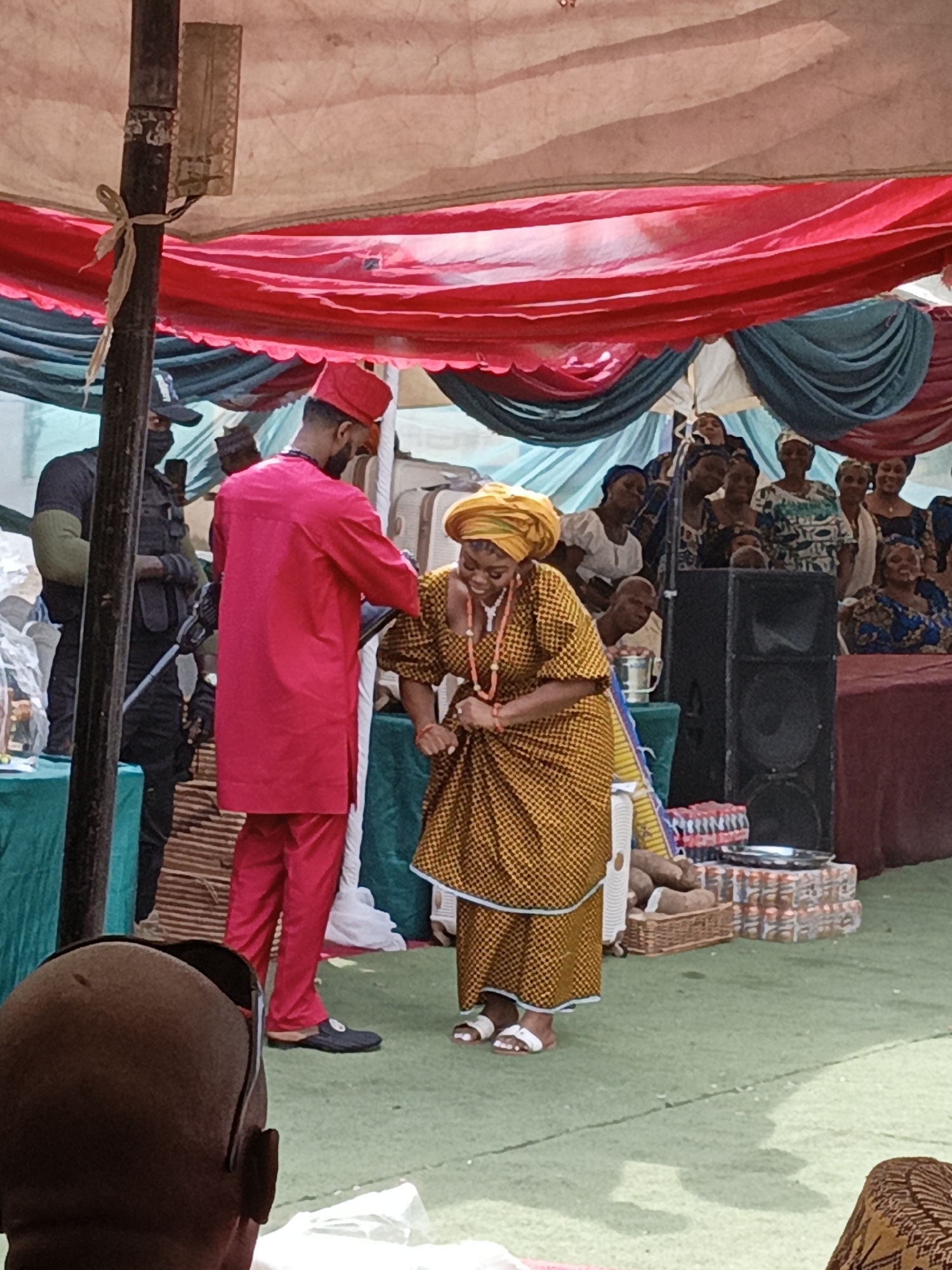 An outdoor cultural celebration unfolds under a large white tent, adorned with flowing red, white, and teal drapes, likely in Sabon Gari West, Nigeria. In the foreground, a man in a vibrant red traditional outfit, including a long top, trousers, and a matching cap, interacts with a woman. The woman, dressed in a golden-yellow patterned traditional wrapper and top, a matching head tie, and an orange beaded necklace, is bending slightly with a wide, joyful smile, holding her dress. The man reaches towards her, suggesting a dance, a blessing, or a traditional ritual common in such festivities.
Behind them, numerous women in diverse traditional attires are seated at tables, observing the event with engaged expressions, some smiling. A man in a black uniform with a face mask and tactical vest stands guard in the left background, adding an element of security to the gathering. The ground is covered with a green carpet or artificial turf. Visible objects include a tall black speaker, stacks of bottled drinks, a basket containing what appear to be yams or tubers, and a silver platter, all hinting at a celebratory feast. The overall atmosphere is festive and joyful, indicating a significant community or family event, possibly a wedding or engagement ceremony, taking place during the daytime.