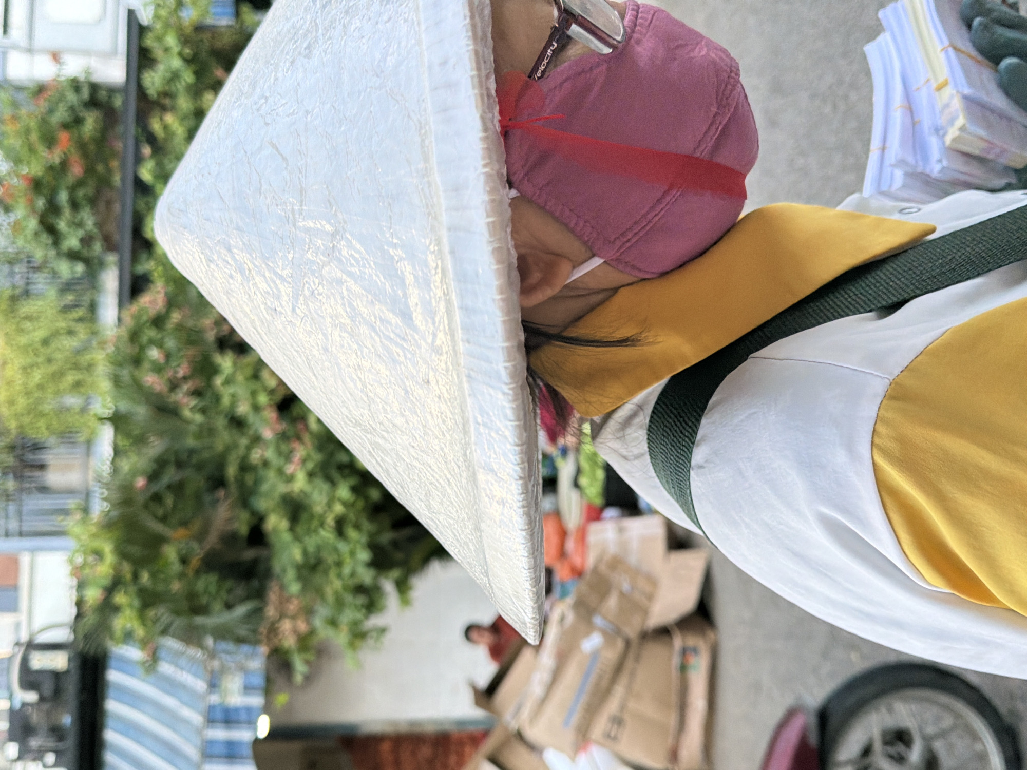 A close-up, waist-up, profile shot shows an individual wearing a conical hat, a pink face mask, and a yellow and white jacket with a green strap. The person is carrying stacks of what appear to be paper currency in their gloved hands. The background is out of focus, but suggests an outdoor market or street scene in Nha Trang, Vietnam. Elements visible include a blue and white striped awning, lush green foliage, a pile of cardboard boxes, and a portion of a motorcycle wheel on the left. The overall lighting suggests daytime.