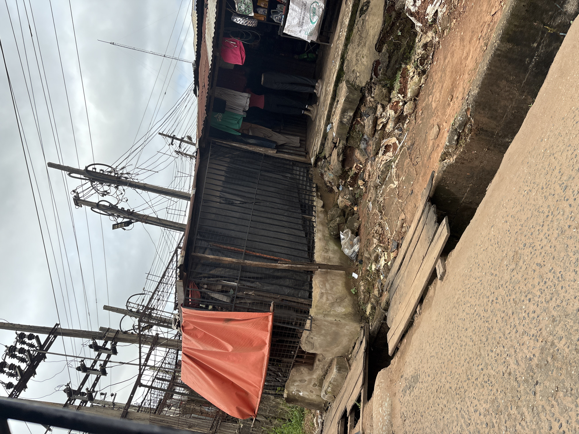 The image depicts a street scene in Nkpor, Nigeria, featuring a roadside shop situated near a street with a drainage ditch. The shop has a corrugated iron roof and is partially covered by an orange tarpaulin. Various items, including clothing, tires, and what appear to be motor oil containers, are displayed for sale. Two mannequins, one in a red top and black pants, and another in a red top and grey pants, are positioned inside the shop. The street in front of the shop is paved with asphalt and shows signs of disrepair, with a wooden plank bridge spanning a section of the drainage ditch. The environment appears cluttered with debris and shows evidence of ongoing construction or damage. Overhead, a complex network of electrical wires and poles dominates the sky.