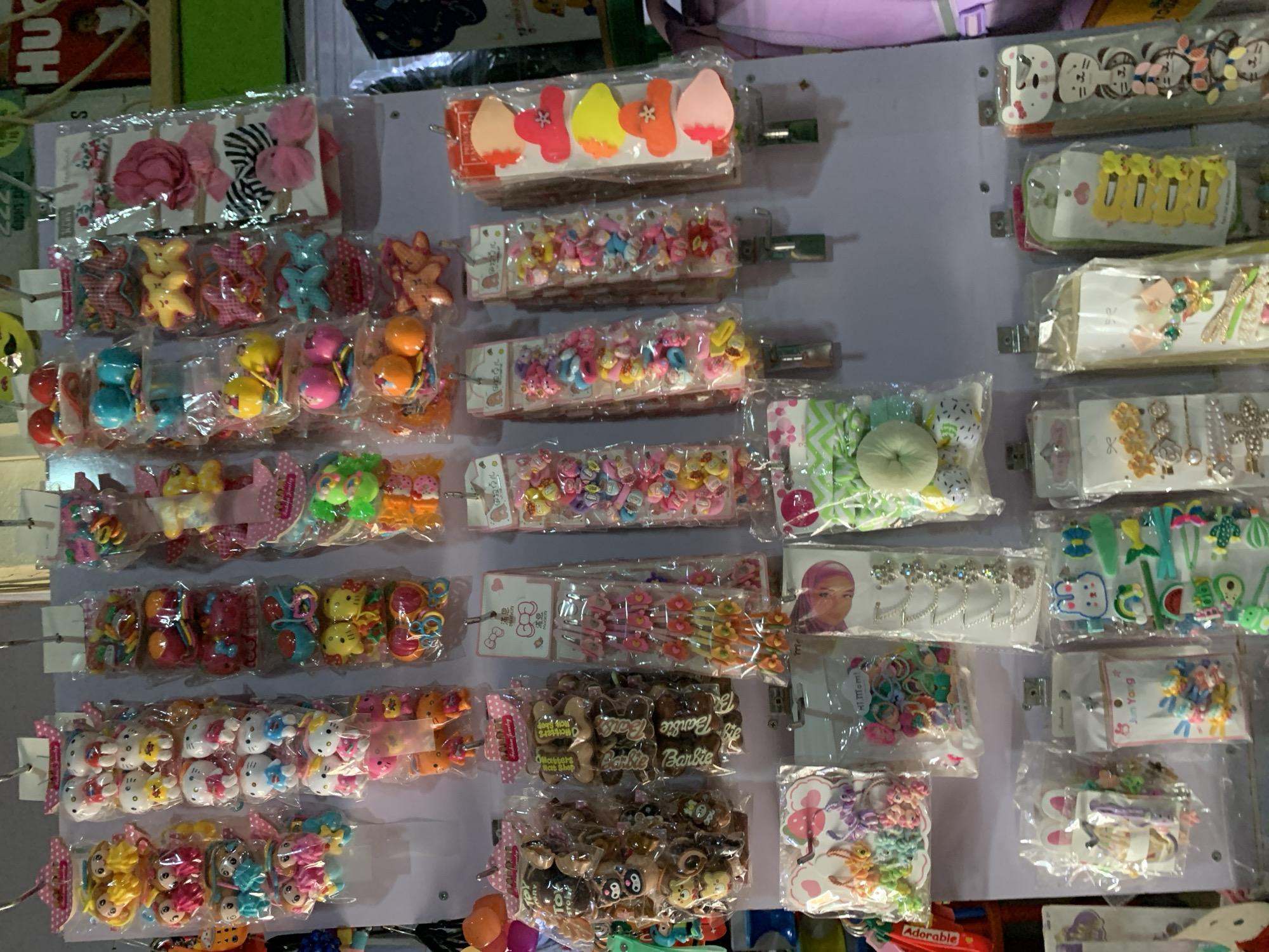 The image shows a display of children's hair accessories mounted on a wall. The display features multiple rows of packaged hair clips and ties, with different designs and colors including cartoon characters like Hello Kitty and Barbie. The accessories are organized on a pale-colored wall, likely in a retail setting. Some printed elements in the image refer to "Barbie" and "Hi.Mom!" and "Jun Yang" as well as "adorable," suggesting brands or slogans. The picture also refers to the city of Jos, Nigeria, based on the location provided.