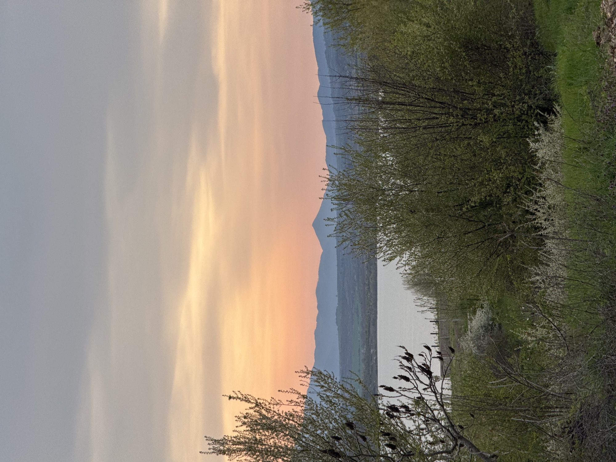 The image depicts a landscape at sunset, likely near a body of water. The sky on the left side is a mix of gray and peach tones, suggesting the setting sun. In the distance, there are hills or mountains appearing as a dark outline against the fading light. The water is in the center of the image with trees and bushes lining the edge, which are more visible on the right side of the image. Some of the trees have begun to bud with new green leaves. The location appears to be Unknown, Unknown.