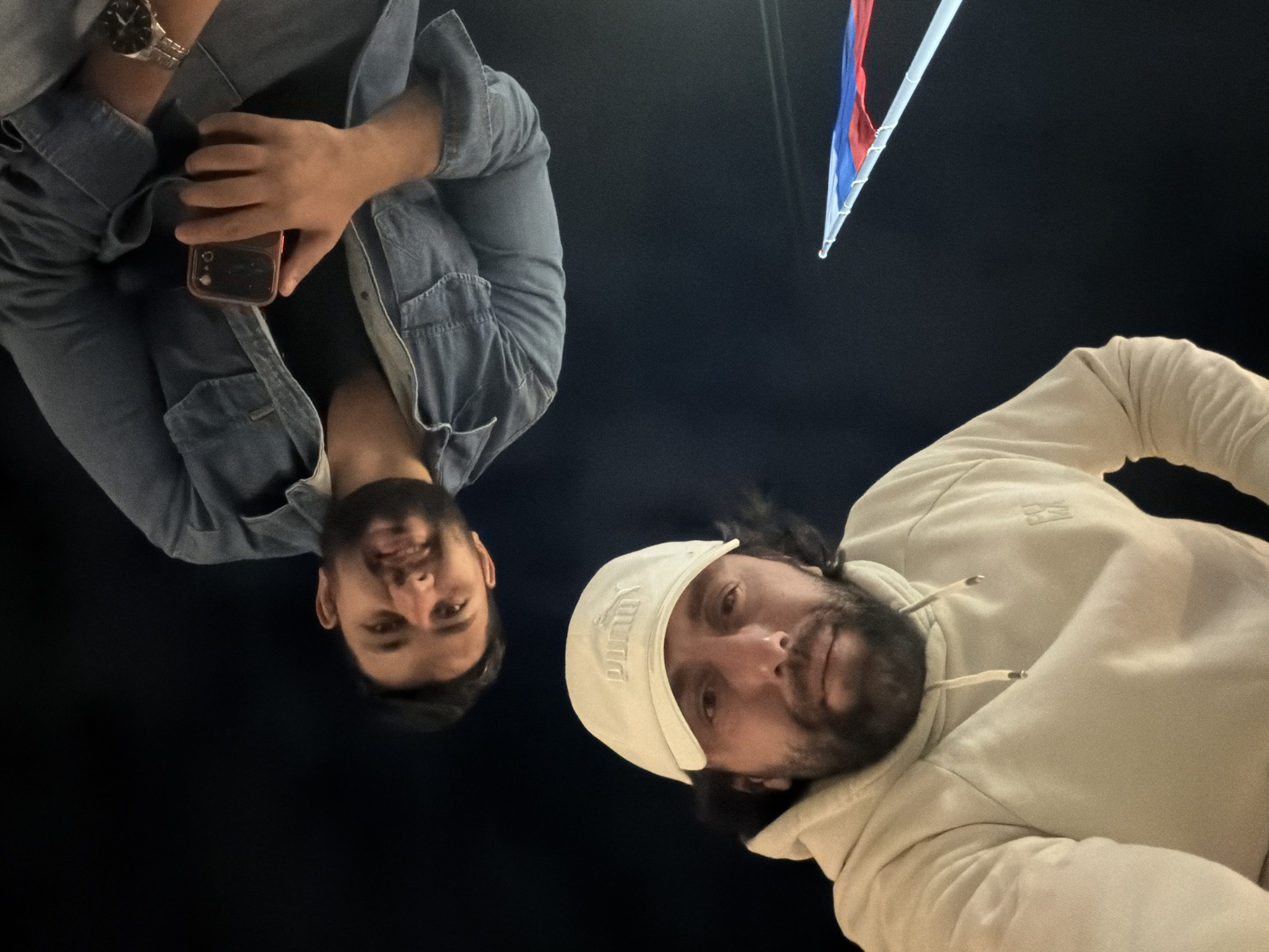 Two men are angled downward at the viewer in an outdoor setting at night. The man on the left is wearing a denim jacket and holding a phone in his left hand. The man on the right is wearing a white baseball cap and a light-colored hoodie. A Russian flag can be seen in the background. The location is Krasnoyarsk, Russia.