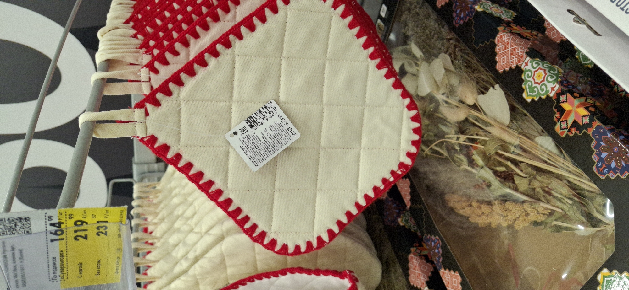 A close-up, eye-level shot shows a rack of quilted kitchen potholders with red zig-zag trim hanging in a store. The potholders are cream-colored with white loops for hanging. A price tag is attached to one of the potholders, showing a price of 164.99 rubles. A barcode and the EAC mark are also visible on the tag. Below the potholders, a clear plastic display case contains a bouquet of dried flowers and decorative items with colorful, geometric patterns. The background of the image is blurred and shows a dark wall with large white lettering, likely part of store signage. The scene appears to be in an indoor retail environment in Telmana, Russia, based on the visible Cyrillic text on the price tag.