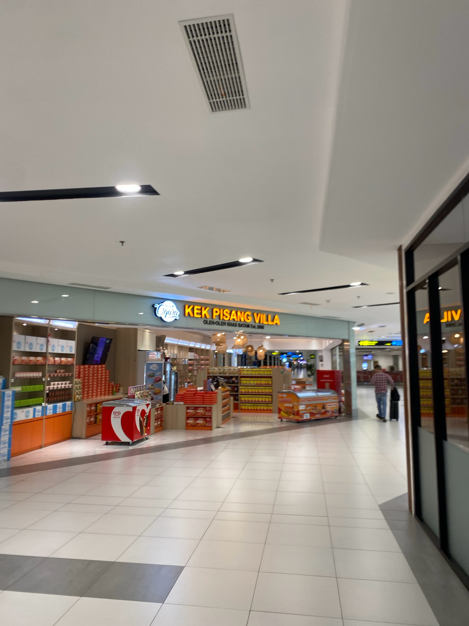 The image depicts the interior of a commercial establishment, likely an airport terminal or a shopping mall, in Batam, Indonesia. The flooring is tiled in a checkerboard pattern of white and grey. Above, the ceiling is white with integrated lighting and a large air vent. In the center of the frame, a shop with a bright orange sign reads "KEK PISANG VILLA" and "OLEH-OLEH KHAS BATAM". The shop displays various products on shelves, including packaged goods and a red Wall's ice cream freezer. To the right of the "KEK PISANG VILLA" shop, another storefront is visible with yellow signage, partially obscured. Further down the corridor, a man in a red plaid shirt and blue jeans is walking away from the camera, pulling a black suitcase. The overall ambiance suggests a retail or transit area with commercial activity.