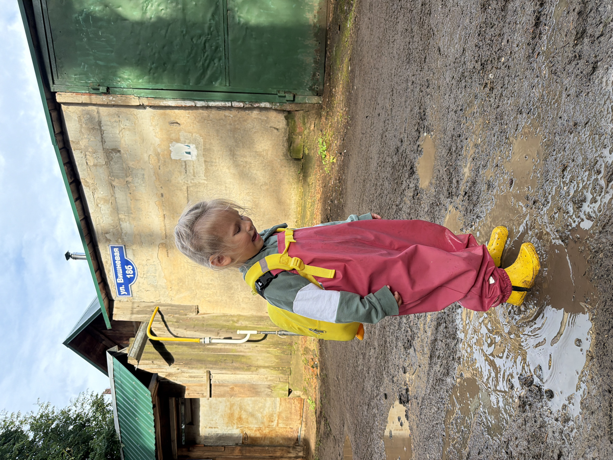 A young girl with blonde hair, wearing a green and white long-sleeved shirt, pink overalls, and yellow rubber boots, stands in a muddy puddle. She has a bright yellow backpack on. The girl is looking down and appears to be smiling. Behind her is a building with a light-colored, textured wall and a large green metal door. A blue street sign that reads "ул. Вишневая 186" (Vishnevaya Street 186) is mounted on the wall. To the left of the building, a structure with a green metal roof is visible, along with a wooden wall and a yellow pipe. The ground is covered in mud and several puddles, indicating a wet environment. The sky above is partly cloudy. The scene appears to be an outdoor residential or industrial area in Chaikovsky, Russia.