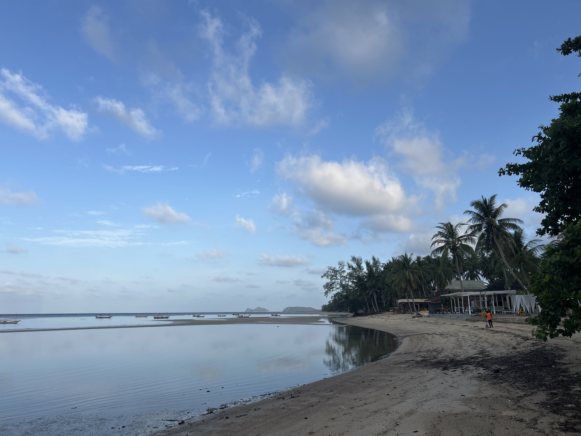 The image depicts a tropical beach scene in the Ko Pha-ngan District of Thailand. The foreground is a sandy beach with some dark debris scattered across it. To the left, calm, shallow water reflects the sky and a line of moored boats. In the mid-ground, a line of palm trees and other vegetation borders the beach. Behind the trees, there are several low-rise structures, possibly bungalows or resort buildings, with white fabric drapes visible. One person, wearing an orange shirt and dark pants, is seen walking on the beach near the buildings. The sky above is blue with scattered white clouds. The overall impression is one of a tranquil beach setting.