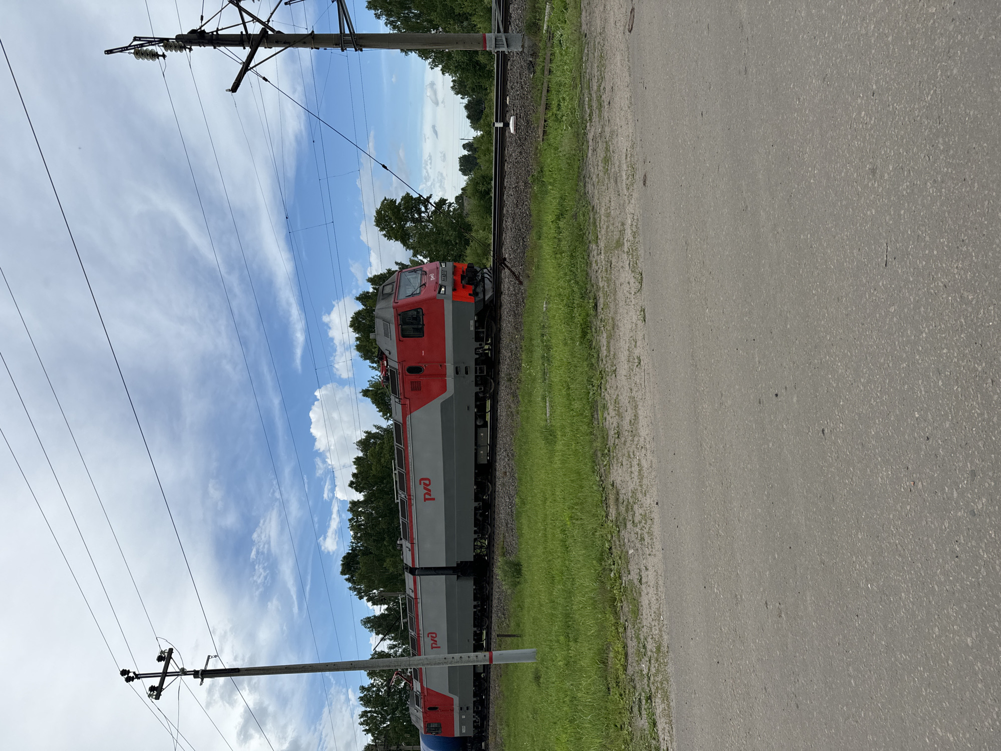 An electric train with red and gray cars, bearing the Russian Railways logo, travels on a railway track in the city of Ponaryrevo, Russia. The train is moving from left to right. Overhead power lines stretch across the frame, supported by utility poles on either side of the tracks. The area is bordered by a grassy embankment to the right of the tracks, and a dirt road or path in the foreground. Lush green trees form a dense line behind the embankment. The sky is a mixture of blue with scattered white clouds. The lighting suggests it is daytime, possibly late morning or early afternoon. There are no people visible in the image.