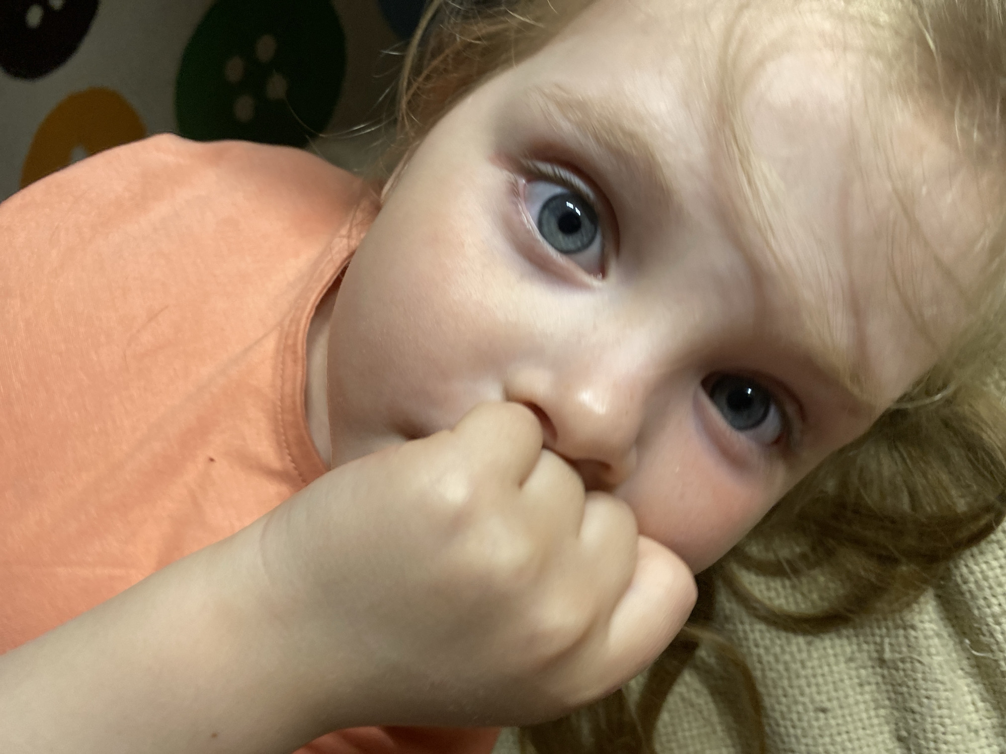 A close-up, slightly angled view captures a young child with blonde hair and blue eyes. The child is wearing an orange shirt. The child's hand is near their mouth, with some fingers partially inserted. The background contains blurry elements of what appears to be a patterned fabric, possibly with button designs. The location is not discernible from the image alone, and there is no information to indicate it is in Votkinsk, Russia.