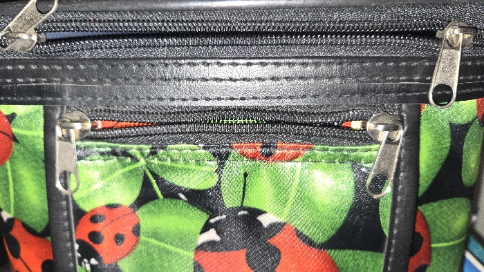 The image showcases a section of a bag featuring two zippers. The top zipper is black, encased in black material. Just below is a strip of black leather, punctuated by stitching. The lower portion of the image displays a colorful fabric adorned with ladybugs and green leaves. Another zipper, this time with an orange interior, rests just above the fabric. Silver zipper pulls are visible on both zippers.