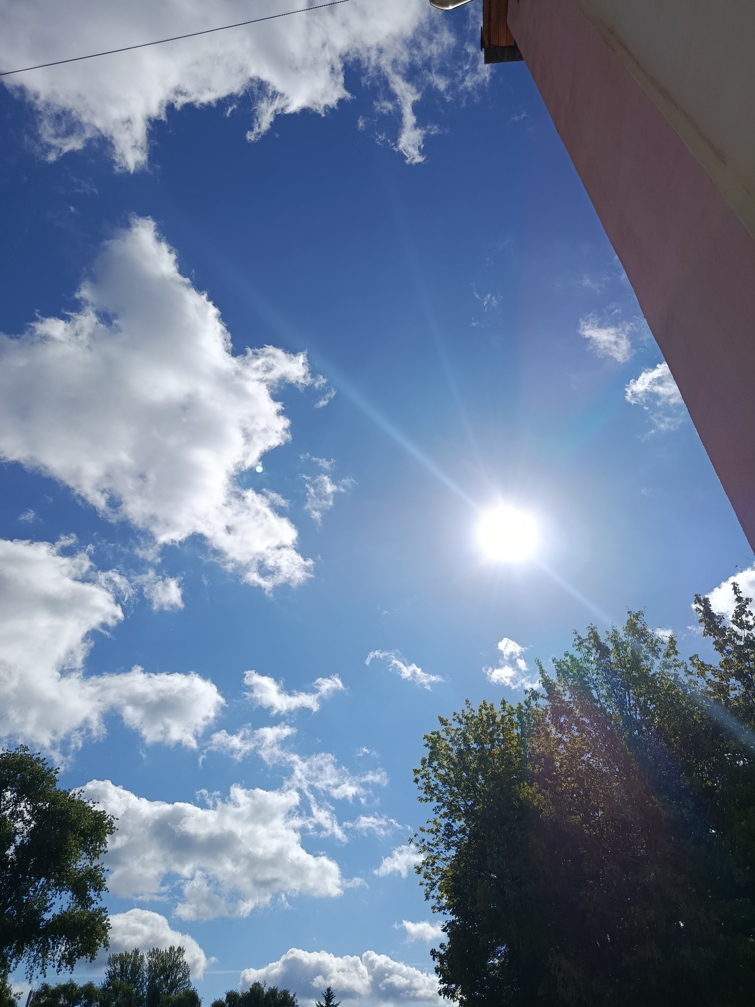 The image depicts a bright, sunny sky with scattered white clouds, as seen from an upward angle. The sun is prominently visible, casting strong rays and lens flare across the scene. In the lower right and left corners, the tops of green trees are visible. A portion of a building, appearing to be a light pinkish-beige color, occupies the upper right quadrant of the image. A thin black wire stretches across the upper left portion of the frame. The location is identified as Svietlahorsk, Belarus, suggesting an outdoor urban or suburban setting on a clear day. There are no people or animals present in the image.