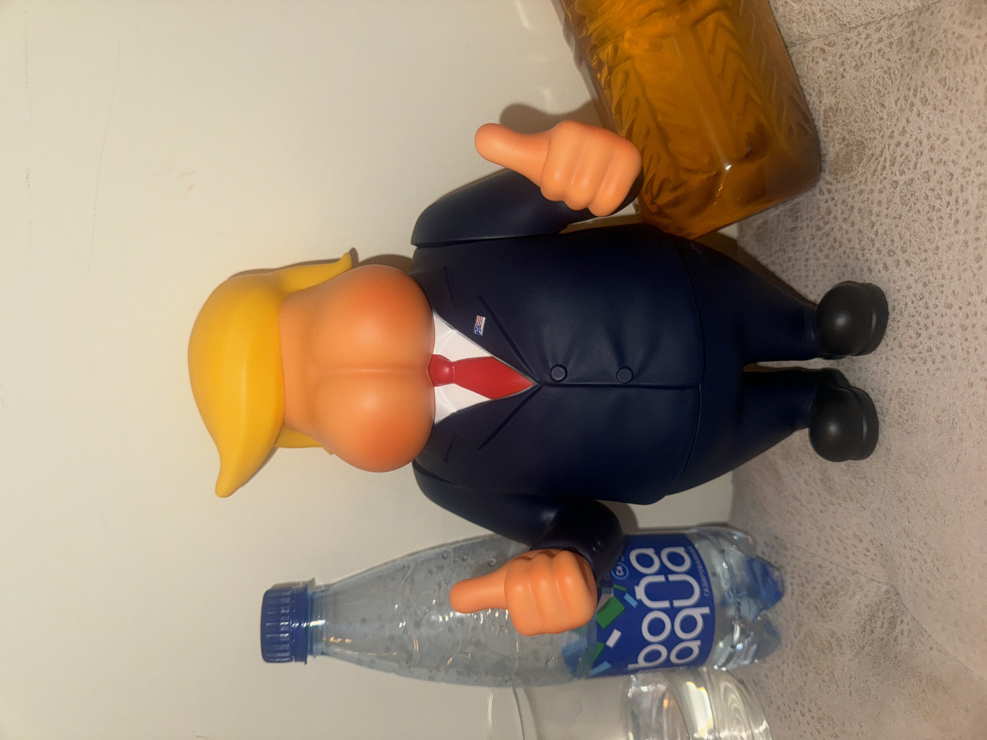 The image shows a plastic figurine of a person with orange skin and yellow hair resembling Donald Trump, wearing a dark blue suit with a red tie. The figurine is positioned between a partially empty plastic water bottle labeled "aqua boba" and a clear plastic bottle with an amber liquid. The background includes a white surface and a light-colored patterned surface. The location in Moscow, Russia, is not apparent in the image.