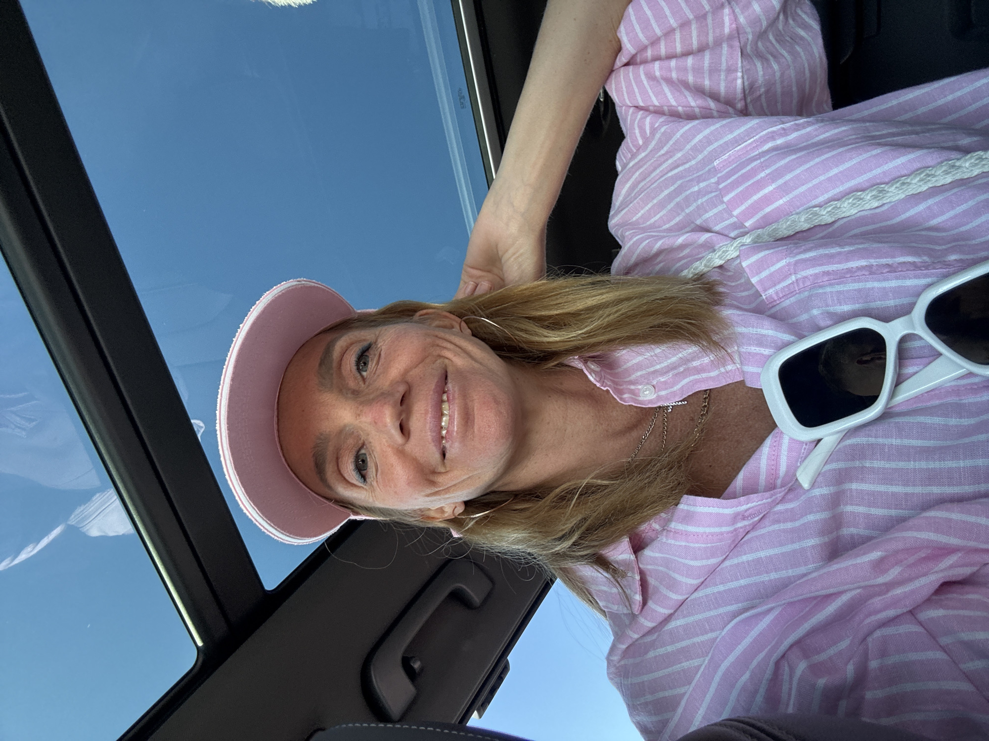 A woman is shown in a selfie inside a car with a glass sunroof offering a view of a clear blue sky. She is wearing a pink and white striped shirt, a pink visor, and white sunglasses, resting on the shirt. She is smiling and has gold hoop earrings. A white braided strap is visible on her shoulder. The interior of the car includes black accents. The location in Kronstadt, Russia is not discernible from the image.
