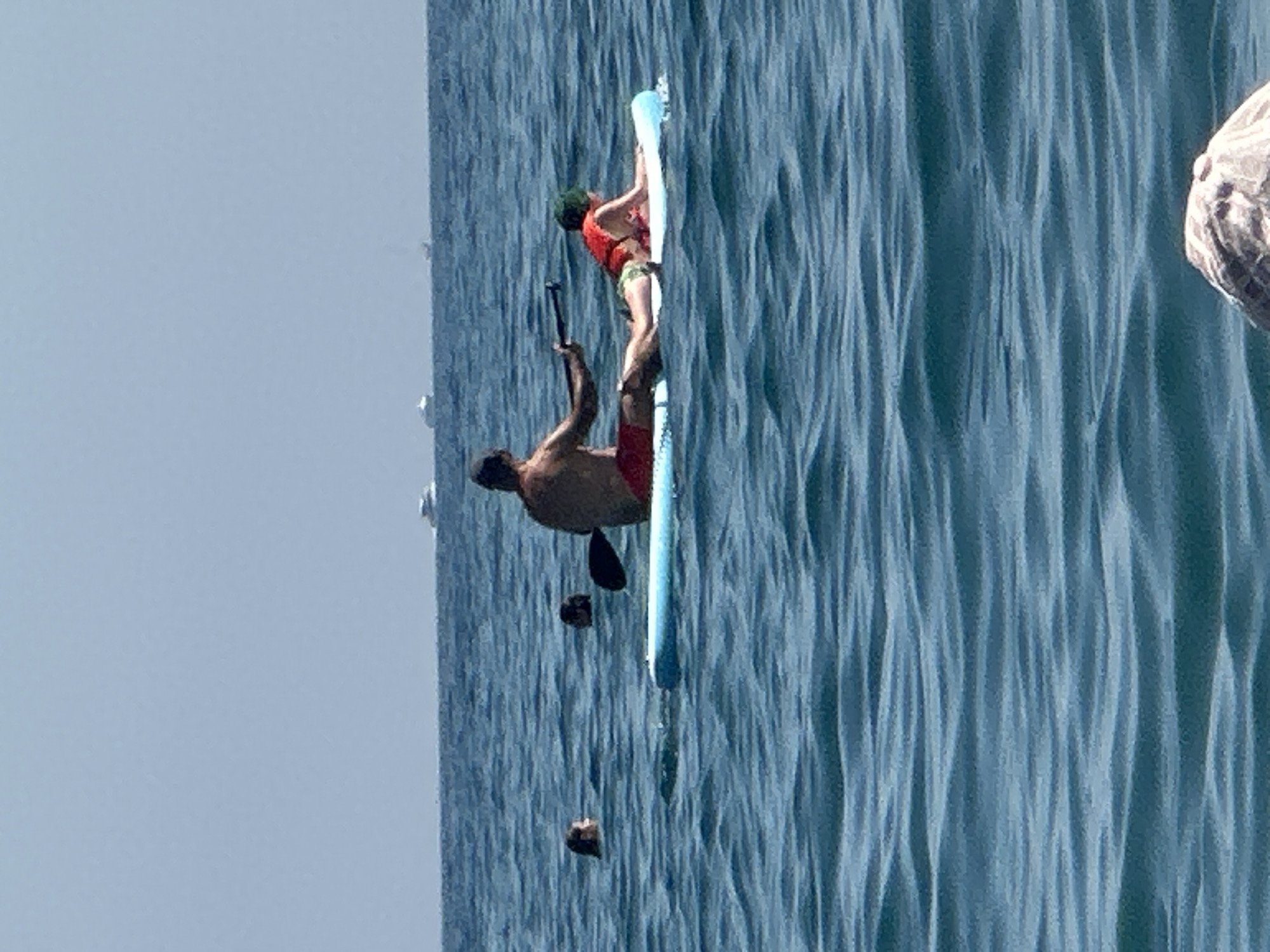 The image captures a bright, sunny day at sea, possibly in or near the city of Boreti, Montenegro. The water is a deep blue with gentle ripples. In the foreground, the edge of a light blue paddleboard is visible. A man, tanned and wearing red swim trunks, is paddling the board while seated. Lying on the front of the paddleboard is a person wearing a red life vest and green shorts, facing forward. Further in the distance, two people are visible in the water, their heads and shoulders above the surface. In the far background, several small boats or ships are spotted on the horizon under a clear, pale blue sky. The scene depicts a recreational activity on a calm day at sea.