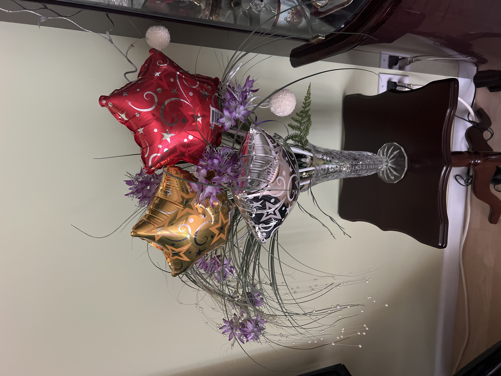 In Farmington Hills, Michigan, a decorative floral arrangement featuring star-shaped balloons is displayed in a crystal vase on a dark wood side table. The arrangement includes three foil star balloons: one gold with a swirl and star pattern, one red with a swirl and star pattern, and one silver and black with a star pattern. These balloons are interspersed with purple artificial flowers and wispy green foliage adorned with small white pearl-like beads. Two fluffy white pom-poms hang from the arrangement. The vase sits on the table, which is positioned against a pale green wall. To the right of the table, a portion of a dark wooden display cabinet is visible, containing various items. Electrical outlets and cords are present on the wall behind the side table. The flooring appears to be wood.