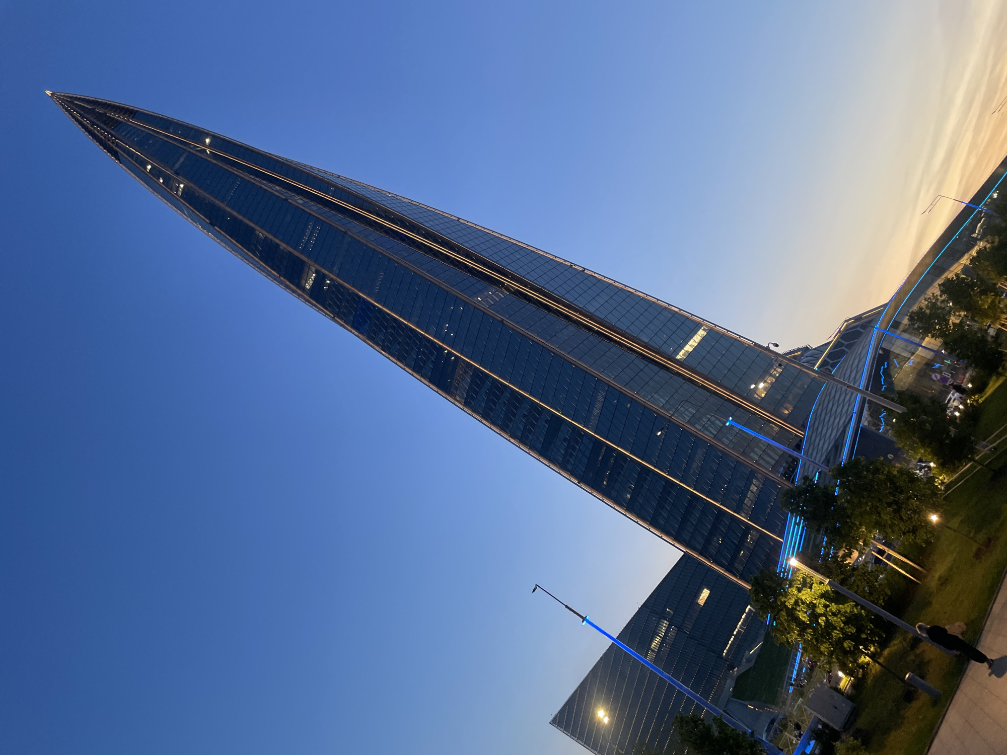 An extreme low-angle, Dutch-tilt shot captures the modern architectural style of a tall, slender skyscraper against a clear blue sky at dusk. The skyscraper's facade is composed of dark glass panels with thin, illuminated horizontal lines running across it. To the left of the main skyscraper, another building with a similar glass facade is visible, also angled upwards. In the foreground, a person in dark clothing walks away from the camera on a paved walkway. Surrounding the walkway are patches of grass with trees and several lampposts. Some of the lampposts and architectural elements of the buildings are illuminated with blue lights, creating a contrasting glow against the darkening sky, which transitions to a pale orange hue near the horizon. The scene is lit by the ambient light of dusk and the artificial lights of the structures.