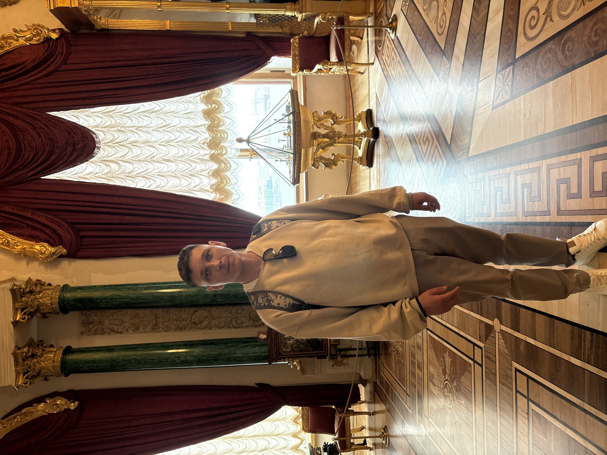A full shot of a man standing in a room. The man is wearing a light beige sweater, light brown pants, and white sneakers. Sunglasses are resting on his chest. He is standing on a parquet floor with geometric designs. Behind him is a large window draped with red curtains and a sheer white curtain. In front of the window is a golden table with a display case. The room is decorated with green marble columns and golden ornaments. This image could be taken in the Winter Palace in Saint Petersburg, Russia.