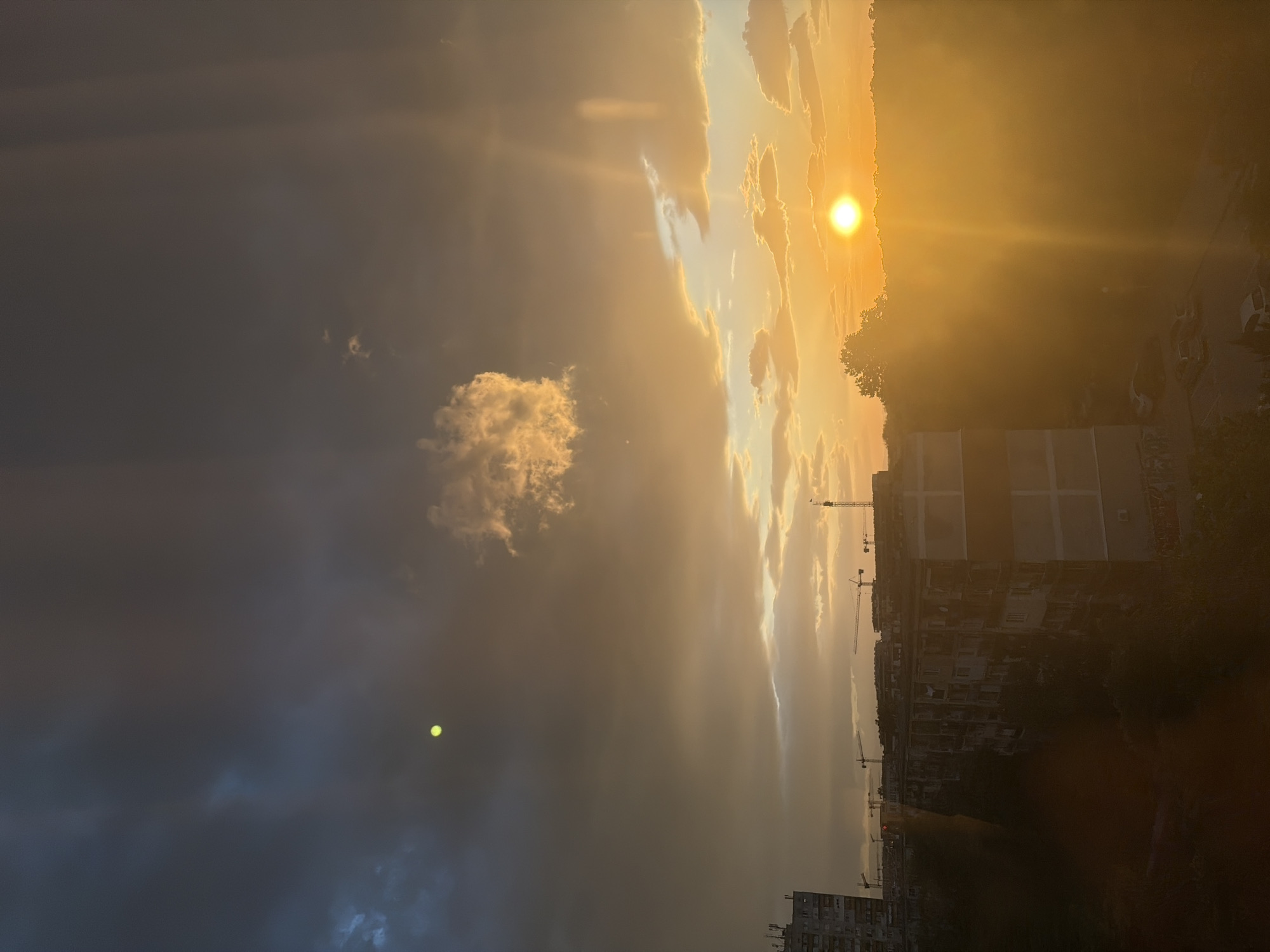 The image shows a view from Plovdiv, Bulgaria, at sunset. The sun is visible through clouds and the air is hazy. In the lower part of the image there are buildings including one with many apartments visible. There are construction cranes in the skyline. To the left is an isolated puffy cloud and in the distance are darker clouds. The photo is oriented vertically.