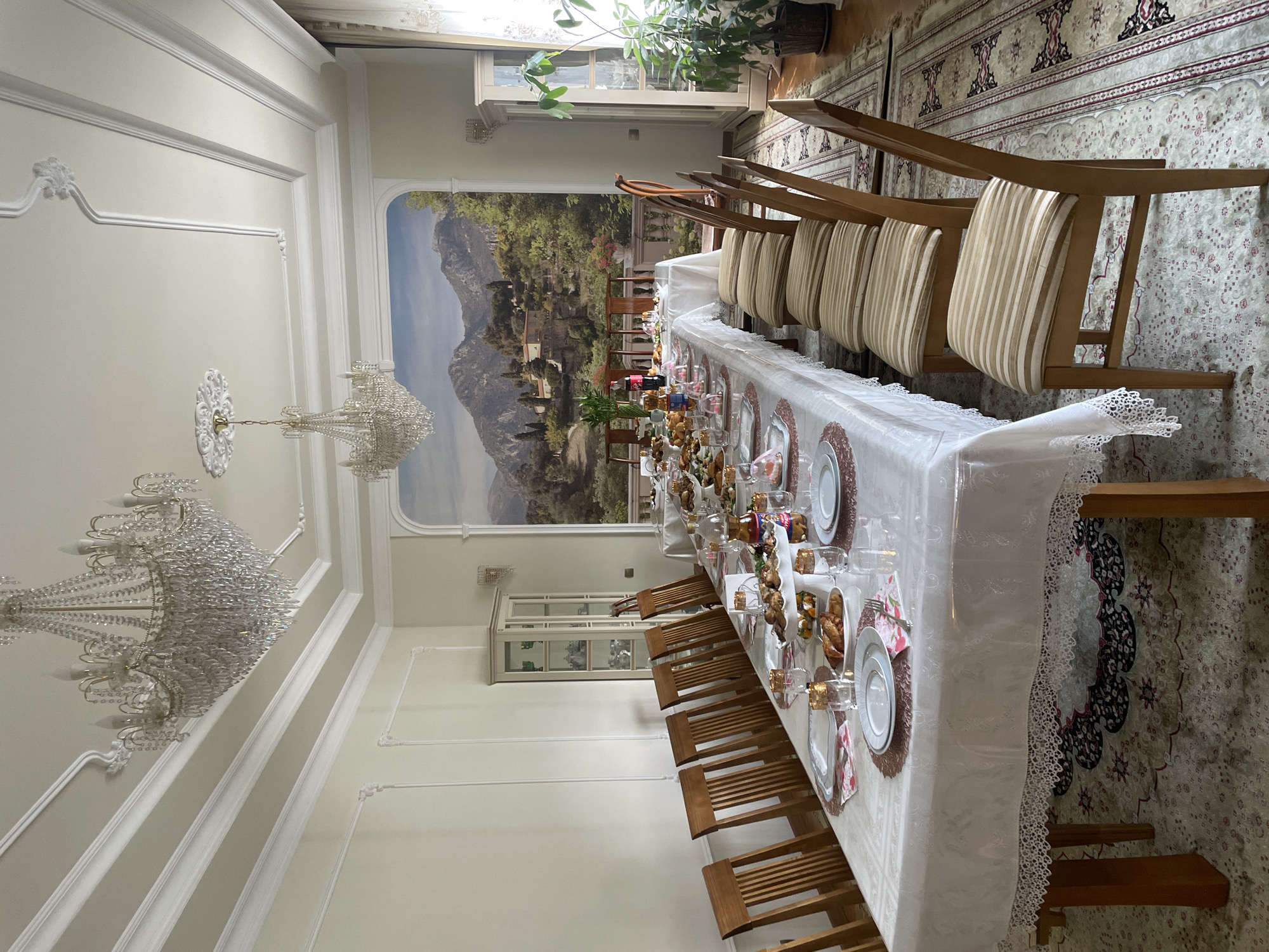 This is a wide-angle, eye-level shot of a formal dining room, possibly in Almaty, Kazakhstan, set for a meal. The room is ornately decorated with white moldings on the ceiling and walls, and two large crystal chandeliers hang from the ceiling. A large mural of a scenic mountain landscape with a villa and trees covers the far wall.
A long, rectangular dining table, draped with a white tablecloth featuring a lace trim, dominates the center of the room. The table is meticulously set with white plates, gold-rimmed glasses, and napkins with floral patterns. Several platters of food, including what appear to be pastries and finger foods, are arranged on the table, along with bottles of drinks.
On the left side of the table, a row of six wooden chairs with slatted backs are visible. On the right side of the table, a different style of wooden chairs with upholstered seats, featuring beige and white stripes, are arranged.
To the left of the table, a tall glass-fronted display cabinet with shelves holding china is present. To the right of the table, a large potted plant adds a touch of greenery to the scene. The floor is covered with a patterned rug. The overall impression is one of elegance and a prepared gathering.