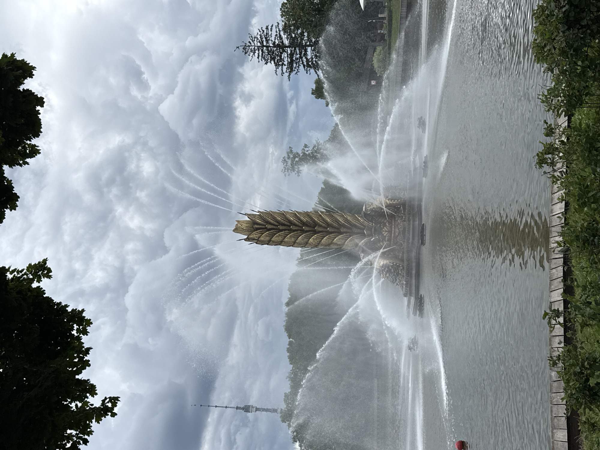 The image shows the Friendship of Nations fountain in Moscow, Russia. The fountain is a large, ornate structure featuring sculptures and numerous water jets. The scene is set in a park or public space with trees and a body of water. The sky is overcast with dramatic clouds, suggesting it is daytime but not necessarily sunny. The fountain is active, with water spraying high into the air, creating a dynamic and energetic display. In the background, a tall tower with a spire can be seen, likely a television tower. The overall mood is one of grandeur and natural beauty, enhanced by the movement of the water and the moody sky. There is no visible text in the image.