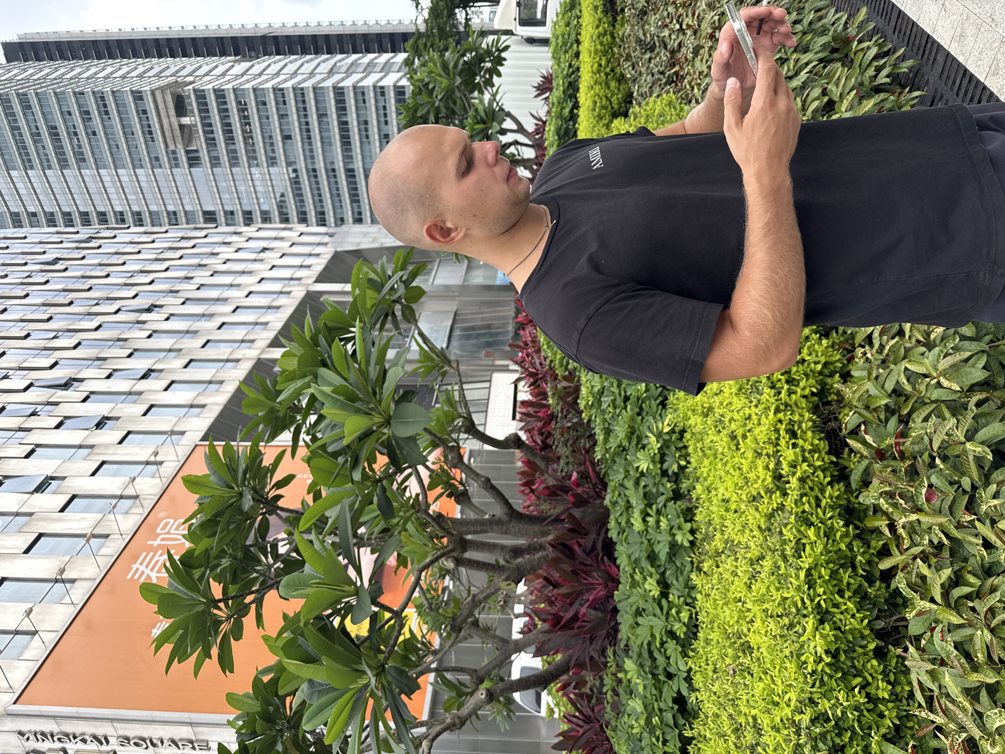 A bald, light-skinned man wearing a black t-shirt and dark shorts stands outdoors in Tianhe District, China, looking down at his phone. The man is positioned in the foreground, slightly to the right of the frame, with a lush green garden and a modern office building behind him. The garden features a variety of plants, including vibrant green shrubs and bushes, as well as red-leafed plants. The building in the background is tall and contemporary, with a facade of windows and orange accents, displaying Chinese characters and the word "SQUARE". The overall scene suggests an urban environment with a focus on landscaping and architecture.