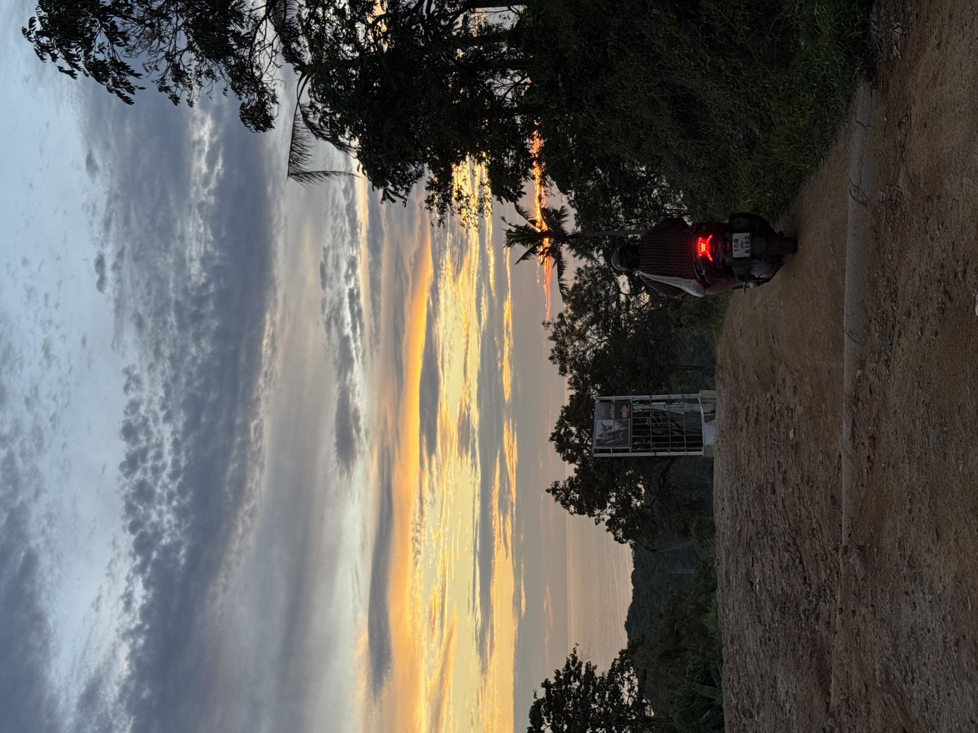A scooter rider on a dirt road faces away from the camera, heading towards a sunset over the ocean in Karon, Thailand. The rider wears a striped shawl and a helmet. The sky is filled with dramatic clouds, with vibrant streaks of orange and yellow near the horizon. Lush green trees line the right side of the road, and foliage is visible in the distance. A metal structure, possibly a signpost, stands on the left side of the road. The road is uneven and appears to be under construction or maintenance. The overall scene captures a serene moment at dusk.
