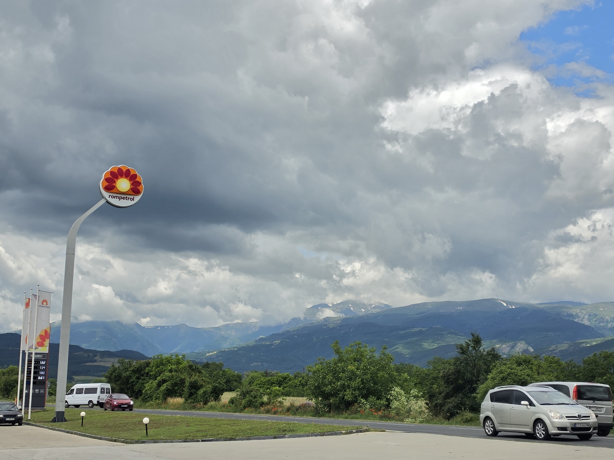 The image captures a view from a roadside service area in Tarnicheni, Bulgaria, under a dramatic, cloudy sky. A prominent Rompetrol gas station sign, featuring a stylized orange and red flower emblem, is visible on the left, mounted on a tall white pole. Below the sign, three tall white flags display pricing information, with visible numbers like "1.62", "1.54", "1.54", "1.80", and "0.67".
Several vehicles are present: a white van is parked to the left of the road, and a red car is driving on the road. Further down the road, a silver minivan is parked, with a portion of a silver SUV visible behind it. The environment includes a paved area, a road, and a grassy verge with scattered red wildflowers. Beyond the immediate roadside, rolling hills covered in lush green trees extend towards a majestic mountain range in the distance. The mountain peaks are partly shrouded in clouds and show patches of snow. The overall scene suggests a stop at a service station during a scenic drive through the Bulgarian countryside.