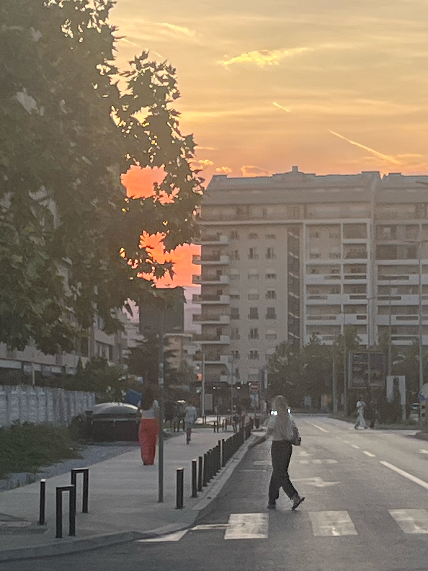 The image captures a street scene at sunset in Podgorica, Montenegro. In the foreground, a pedestrian crosswalk is visible on the right side of the road, with a person walking across it. The person is wearing dark pants and a light-colored top, carrying a bag over their shoulder. The street is lined with bollards on the sidewalk. To the left of the sidewalk, a large tree with green foliage partially obscures the view of the sky and the buildings behind it. The sunset is casting a vibrant orange and yellow glow, visible through the gaps in the tree leaves. Beyond the tree and sidewalk, there are several multi-story apartment buildings with a repetitive architectural style. Further down the street, more pedestrians and a vehicle can be seen. The sky is filled with soft clouds and has the distinct appearance of a setting sun, with contrails from an aircraft visible. The overall impression is of a typical urban evening scene in Podgorica.