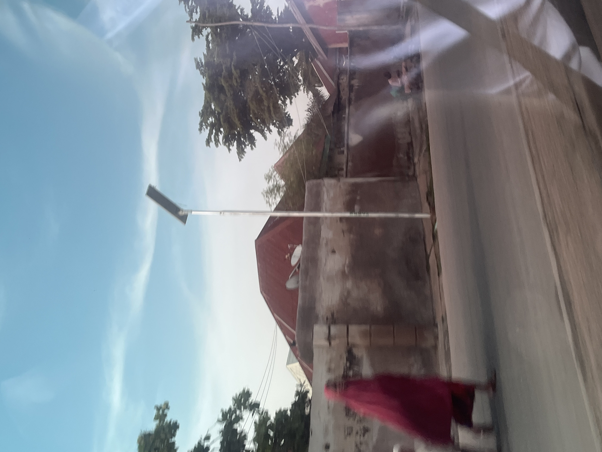 The image depicts an outdoor scene in Jalingo, Nigeria, oriented vertically. In the foreground, a person wearing a maroon garment is partially visible, their back turned towards the camera. The person appears to be standing or walking alongside a textured wall. In the background, a street is visible, with a few buildings on the right side. One building has a red, sloped roof and a lighter-colored upper section. Another structure is a tall, light-colored wall with some visible tiling. A street lamp with a solar panel is positioned on the left side of the frame, extending into the sky. Trees with dense foliage are present in the upper left and lower right portions of the image. The sky is a light blue with wispy clouds, suggesting it is daytime, likely late afternoon or early morning due to the soft lighting. The overall impression is a view of a residential or semi-urban area. There is no visible text in the image.