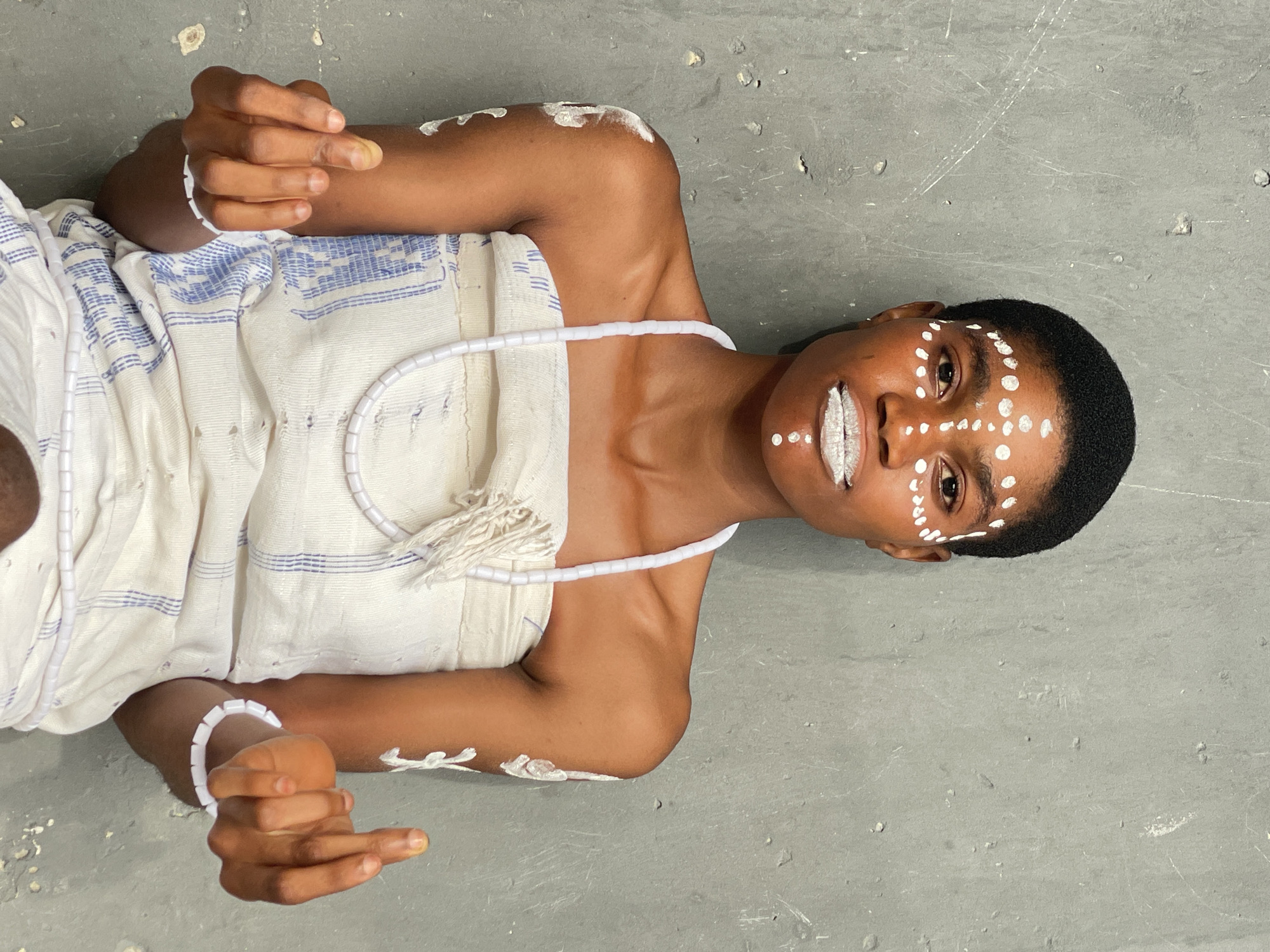 A person lies on a gray surface. The person is Black and adorned with white markings on their face, shoulders, and arms. They wear a white cloth garment with blue stripes and a white beaded necklace and bracelet. The location is not discernible from the image.