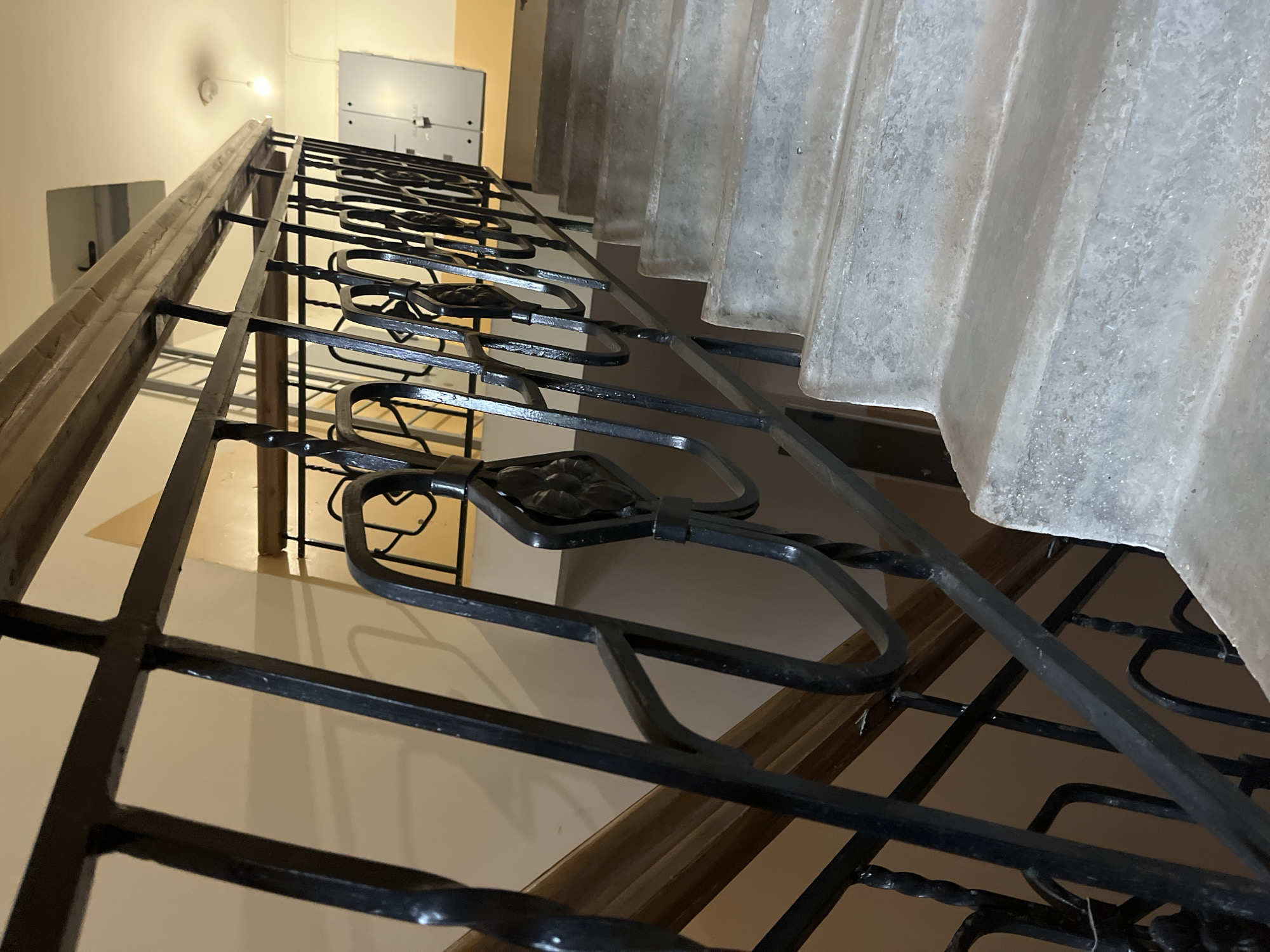 The image presents an interior view of a stairwell, captured from a low angle looking upwards. A prominent, dark wrought iron railing with intricate decorative scrollwork and floral motifs runs along the left side and center of the frame, leading the eye upwards. The top of the railing is capped with a weathered wooden handrail.
To the right, a wide concrete staircase ascends, displaying a rough, textured, and somewhat worn surface. The walls are painted in a light, neutral tone, possibly beige or cream, with a darker wooden baseboard or trim running along them. In the upper background, two light grey electrical or utility boxes are mounted on a wall. A single, exposed light bulb fixture is visible on the ceiling or upper wall, providing artificial illumination. No people are present, and there is no visible text. The overall scene suggests an aged or utilitarian environment within a building.