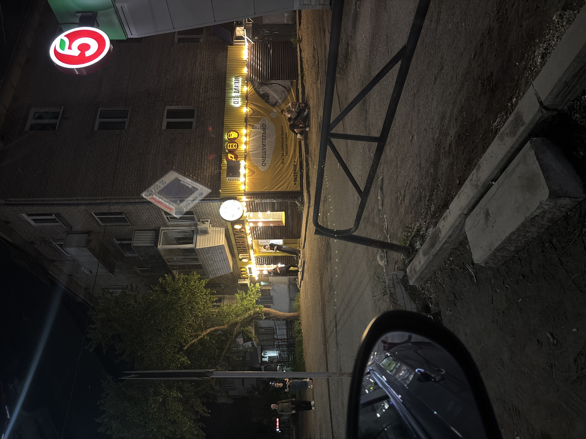 The image captures an urban scene at night in what appears to be the city of Chaikovsky, Russia. Visible in the upper left is a lit circular sign with the number "5" in red and green. Adjacent to it is a multi-story brick building. Next to the brick building, there is a banner displaying the words "СЕТЬ МАГАЗИНОВ ОЧЕШУИТЕЛЬНО!". The banner includes the text "ВСЕГДА ШИРОКИЙ ВЫБОР СВЕЖЕЙ РЫБКИ" and features icons representing beverages, food and fish, alongside the word "Пенное".
To the right of the building is a black metallic structure. Further down the street, there's a scattering of people, some standing and others sitting. Trees are also present in the mid-ground. In the foreground, a car side mirror reflects elements of the cityscape.