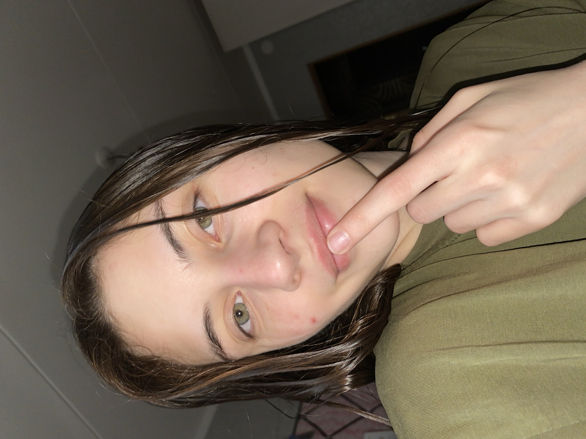 A close-up, angled portrait shows a person with fair skin and dark brown, wet hair. They have green eyes and are touching their upper lip with the index finger of their right hand. The person is wearing a green shirt. The background is dimly lit, showing a portion of a wall and a dark object. The location is not discernible as Votkinsk, Russia.