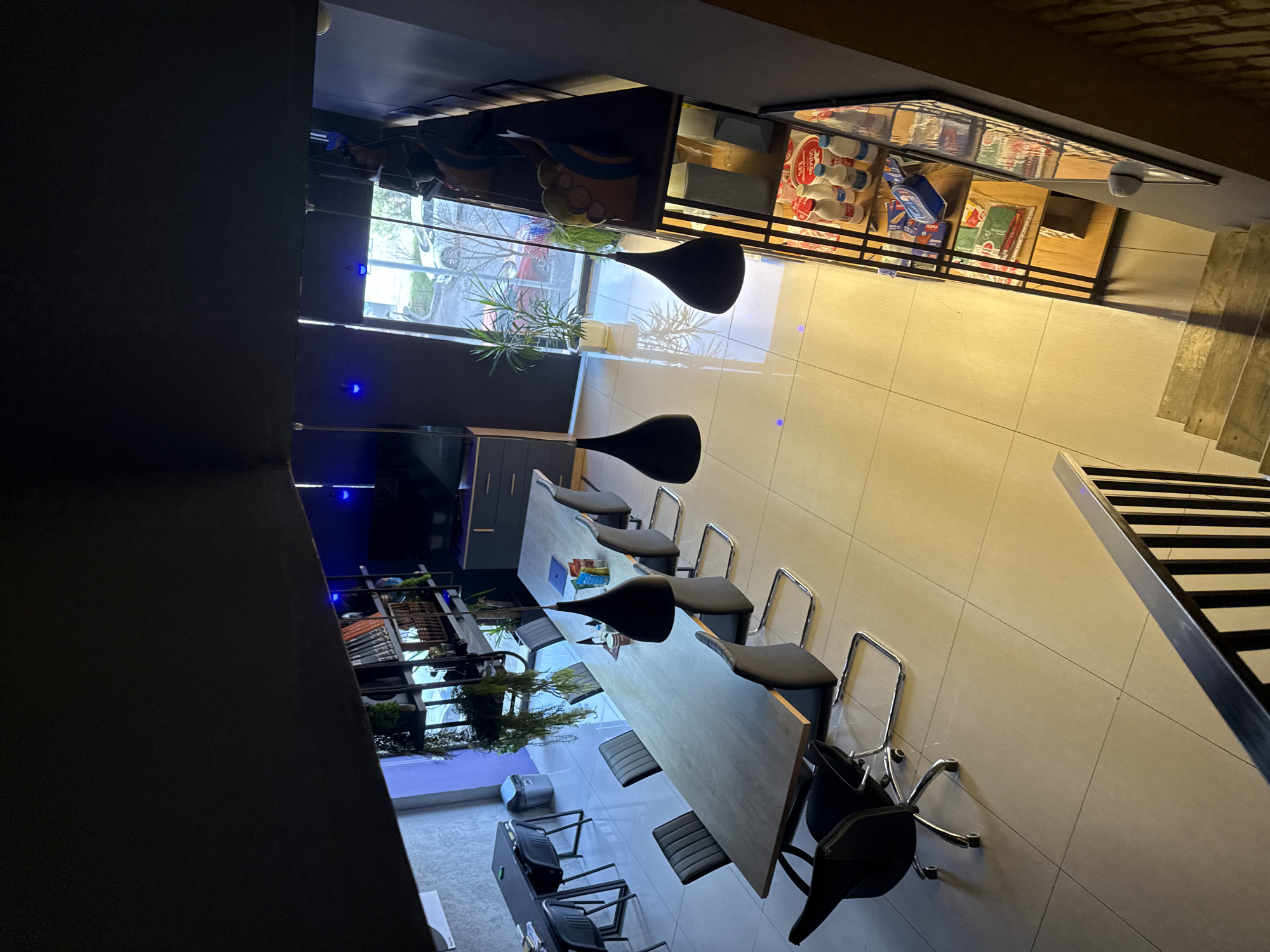 The image depicts an interior view of a commercial space, potentially a restaurant or cafe. The location is likely in Yerevan, Armenia, although this is not explicitly indicated within the image. The room features a long, wooden table surrounded by modern office chairs. There are decorative plants near a large window, suggesting an effort to bring nature indoors. A display case above the seating area contains various products, including beverages, which points to the commercial nature of the space. The floor is tiled, and stairs are visible in the corner, suggesting a multi-level establishment. The room is well-lit and appears to be clean and organized.