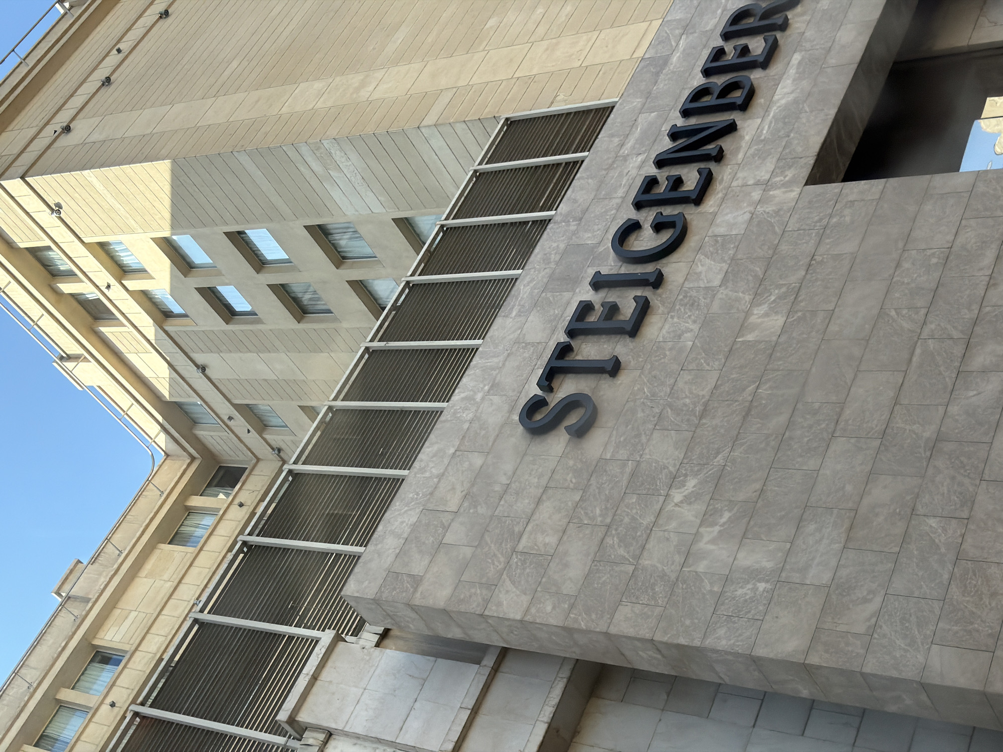 The image shows the exterior of the Steigenberger El Tahrir hotel, a six-story building located in Cairo, Egypt. The hotel features a modern architectural design with a light beige stone facade. The building has numerous windows, many of which are covered by horizontal louvers. The name "STEIGENBERGER" is prominently displayed in large black lettering on the lower portion of the building, above the entrance. The sky is clear and blue, indicating a sunny day. The image is taken from a low angle, looking up at the hotel.