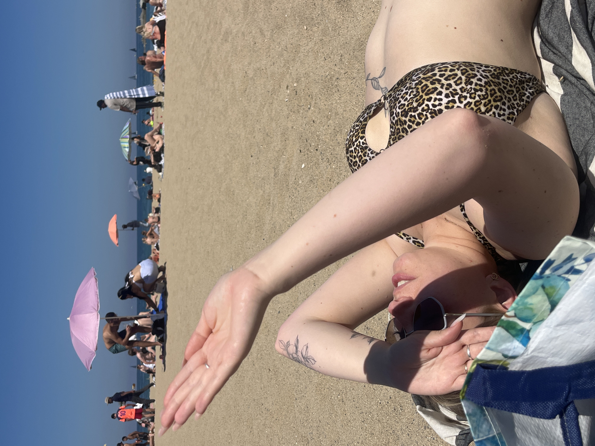 A woman lies on a sandy beach, seemingly enjoying the sun. She is wearing a leopard-print bikini. Her arms are raised, shielding her face. She is also wearing sunglasses. She is on a beach filled with people, some of whom are under umbrellas. It's a sunny day. The location is Barceloneta Beach in Barcelona, Spain.