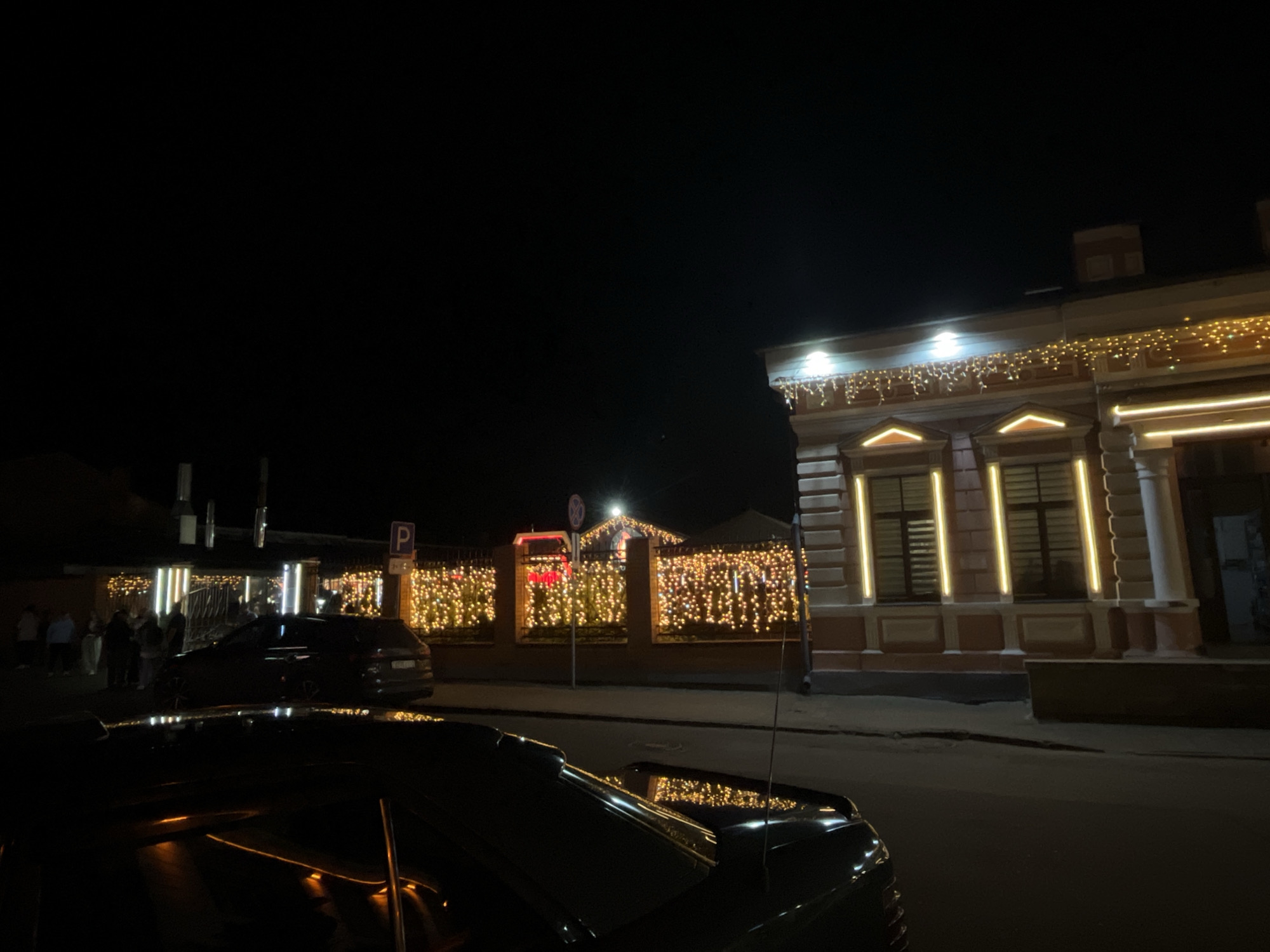 A night-time outdoor shot captures a street scene in Vitsebsk, Belarus, illuminated by festive decorations. On the right, a building with classical architectural elements is adorned with strings of warm, yellow lights along its facade and roofline. To the left, a fence is also covered in a dense curtain of similar twinkling lights, creating a vibrant glow. Several people are visible on a sidewalk in front of this decorated fence. Parked cars are present on the street in the foreground, with their reflections visible on their surfaces. The overall atmosphere suggests a celebration or holiday event.