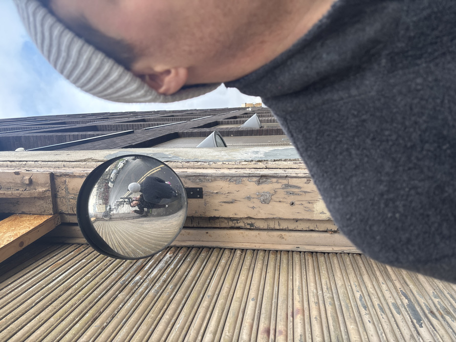 A first-person perspective photograph captures a street scene in the City of London, United Kingdom, on an overcast day. The right side of the frame shows the right ear, jawline, and shoulder of an individual wearing a light grey ribbed beanie and a dark grey, textured jacket. To the left, a circular convex security mirror is affixed to a worn, pale-colored building pillar, which exhibits peeling paint and exposed wooden elements, including a raw timber beam at the top. Adjacent to the pillar on the left is a closed, horizontal-ribbed metal roller shutter. To the right of the pillar, a dark brick building facade with several light-colored, angled awnings or fixtures is visible. The convex mirror's reflection reveals a wet street scene: two individuals are observed, one with their back to the reflection's foreground (wearing a grey beanie and dark jacket, consistent with the person holding the camera), and another individual facing them, gesturing with their right hand. The reflected street is lined with black bollards and features parked cars on the right side. The sky in the reflection is consistent with the visible cloudy sky.