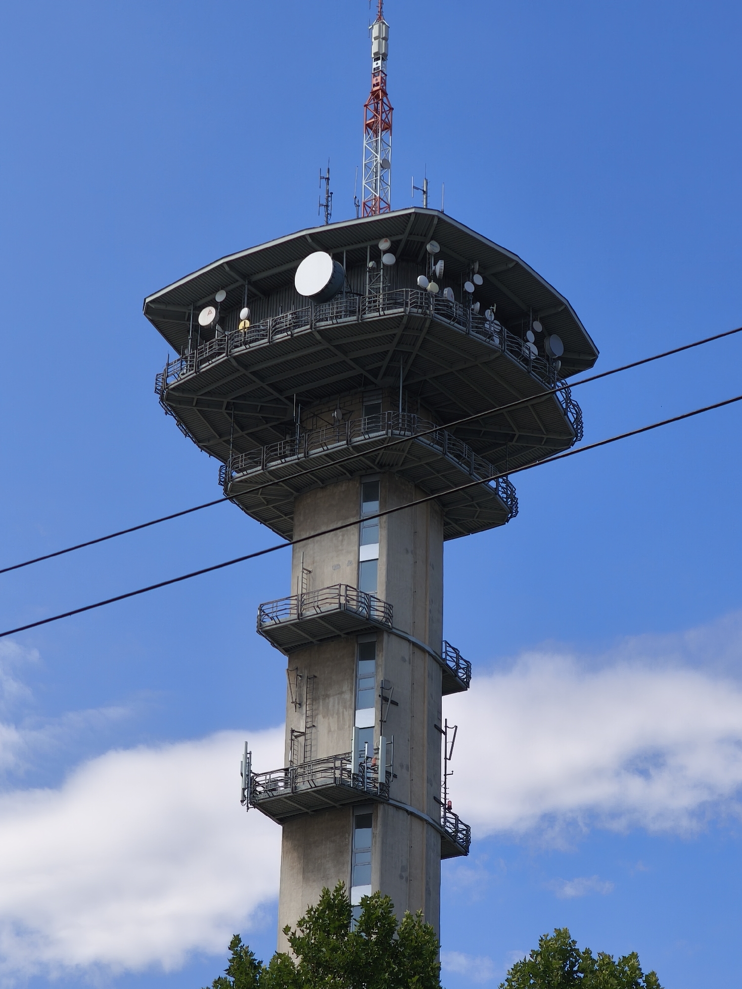 The image shows a tall concrete transmission tower with a wider, multi-tiered structure at the top, adorned with various antennas, dishes, and a red and white lattice mast at its peak. The tower is set against a bright blue sky with scattered white clouds. Two dark power lines stretch horizontally across the frame, obscuring portions of the sky and the tower. The tower has several external staircases and balconies with railings. Lush green trees are visible at the base of the tower. This is the Dubany transmitter in Pardubice, Czechia.