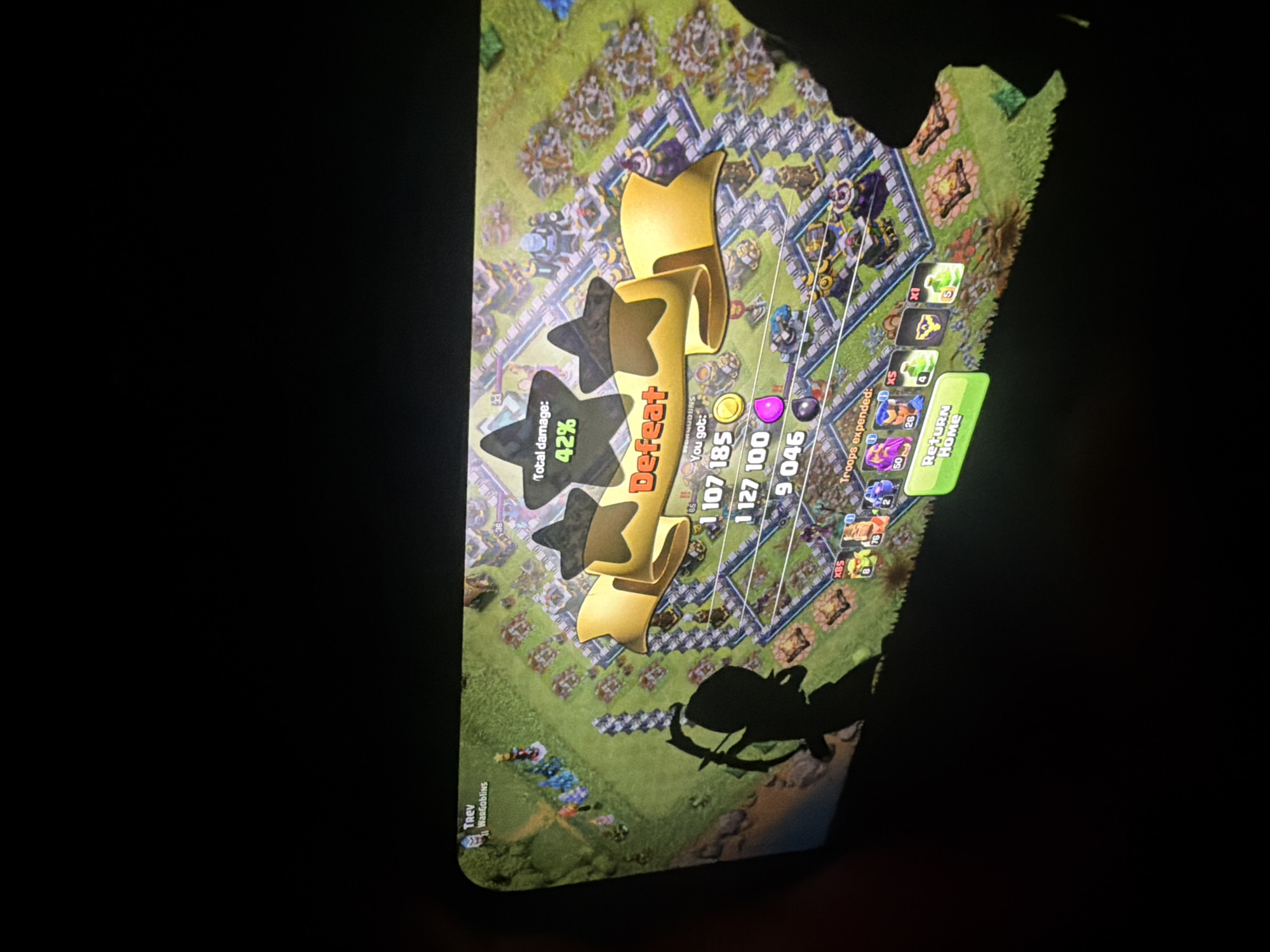 The image displays a mobile device screen showing the post-battle results from the game Clash of Clans. The primary focus is the game's interface, which is partially obscured by a dark surrounding.
The screen indicates a "Defeat" with "Total damage: 42%," suggesting a failed attack in the game. A golden banner with three blacked-out stars highlights this outcome. Below the "Defeat" banner, text states "You got:" followed by resource amounts: "1 107 185" (gold), "1 127 100" (elixir), and "9 046" (dark elixir). Further down, "Troops expended:" is visible, alongside icons and numbers indicating various units used in the battle, such as "x85," "50," and "26." A green button at the bottom right prompts "Return Home."
On the left side of the screen, the player's name "TREV" and clan name "WarGoblins" are visible. The game's setting, a fortified village with various buildings and defenses, is rendered in detail. A prominent shadow of what appears to be an archer or a person holding a bow and arrow is cast across the lower right portion of the game screen, suggesting an external object or a player's hand interacting with the device. The dark background surrounding the bright game screen implies that the image was captured in a dimly lit environment.