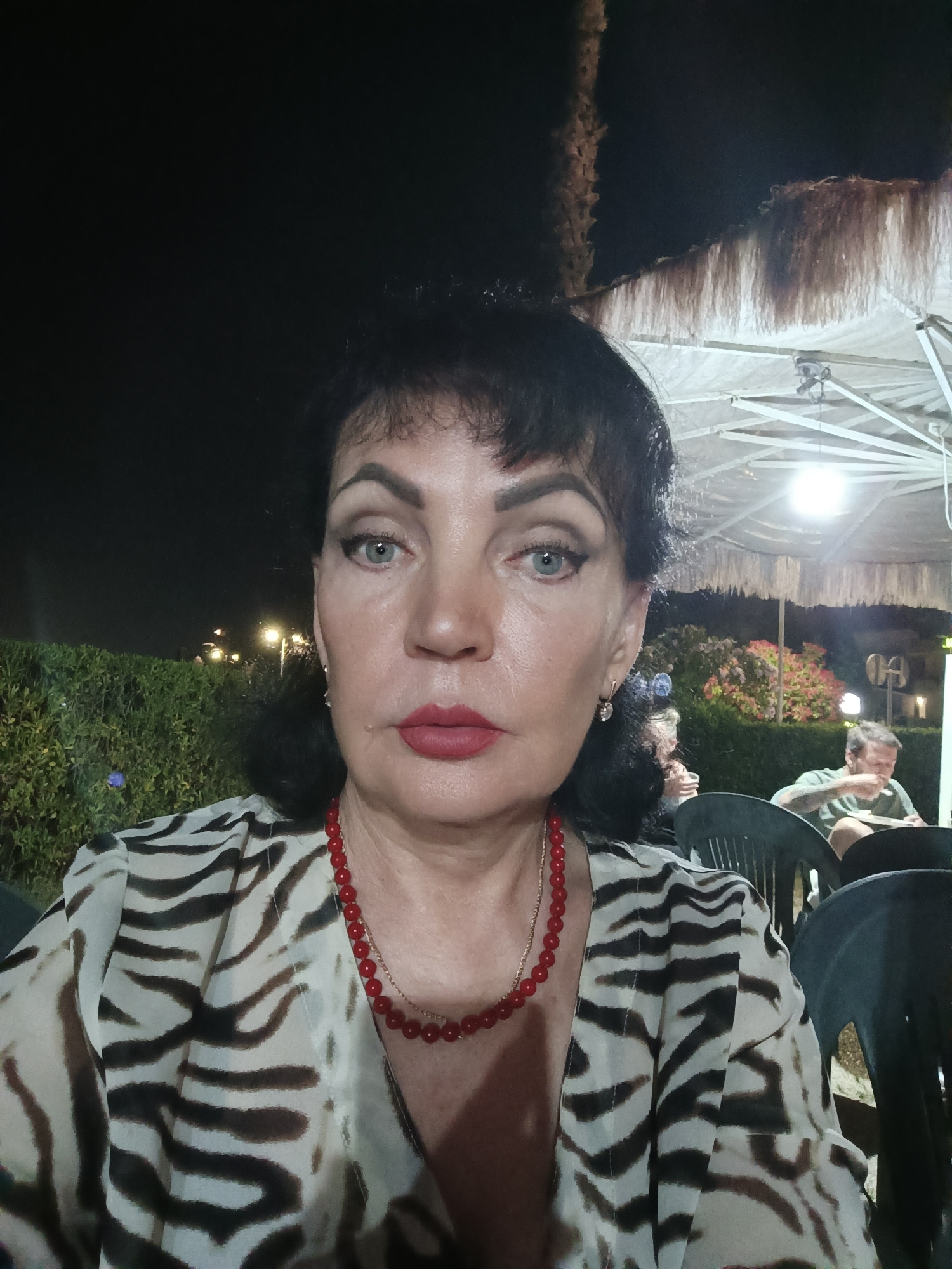 A woman with dark hair and fair skin, wearing a zebra-print blouse and a red beaded necklace, poses for a selfie. Her makeup includes red lipstick and winged eyeliner. In the background, under a thatched umbrella, a man sits at a table, eating. The setting appears to be an outdoor seating area at night, with plants and lights visible. The location is not specified as Giulianova, Italy.