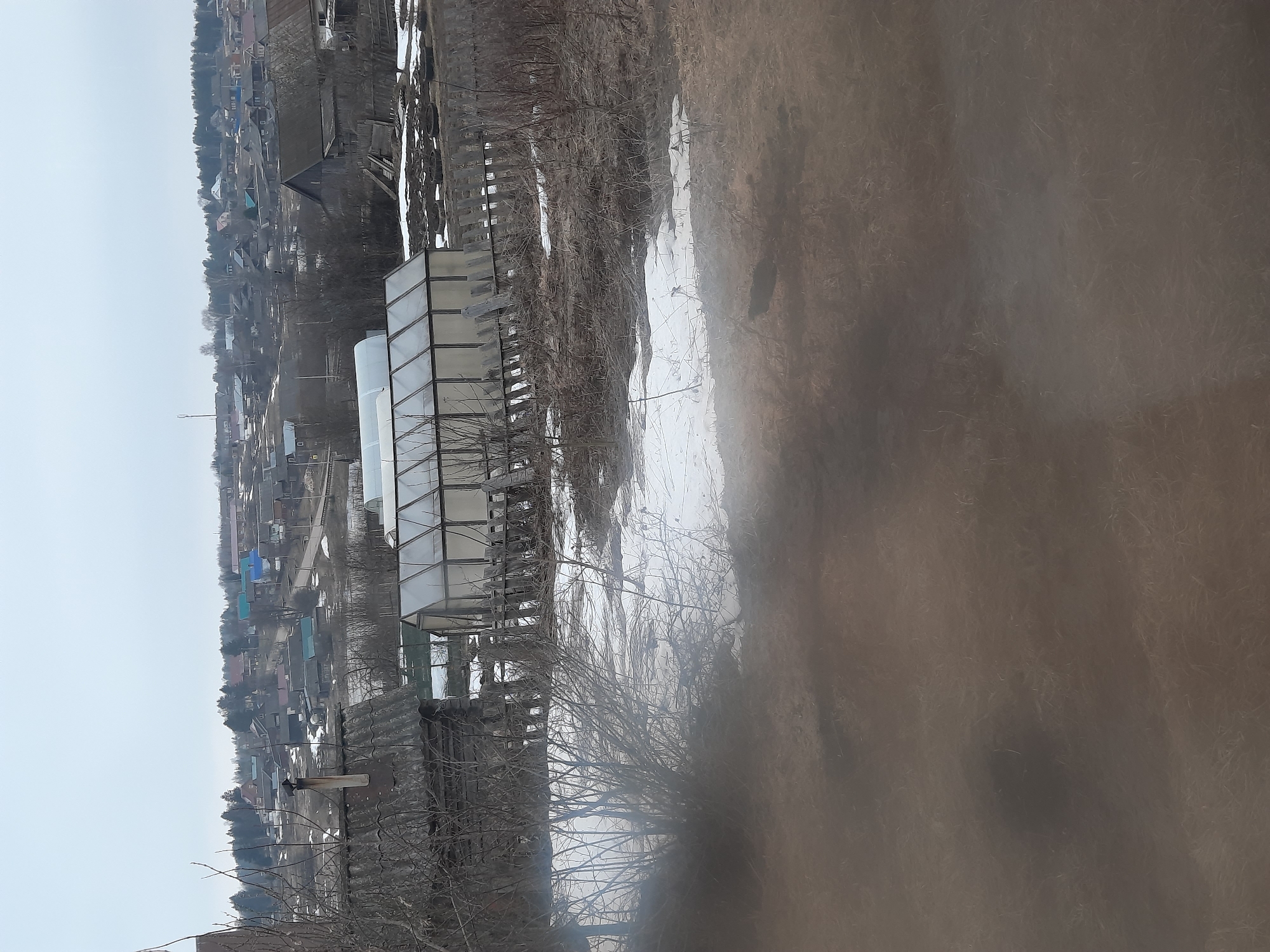The image depicts a view in or near Krasnogorskoye, Russia. In the foreground, there is a bare bush or small tree. Behind it, there appears to be a greenhouse structure. Patches of snow are visible on the ground. Further in the background, there's a settlement with houses and some trees, possibly indicating the edge of the town. The sky is overcast, suggesting a cloudy day. The immediate foreground is somewhat blurry, perhaps due to condensation or a smudge on the lens.