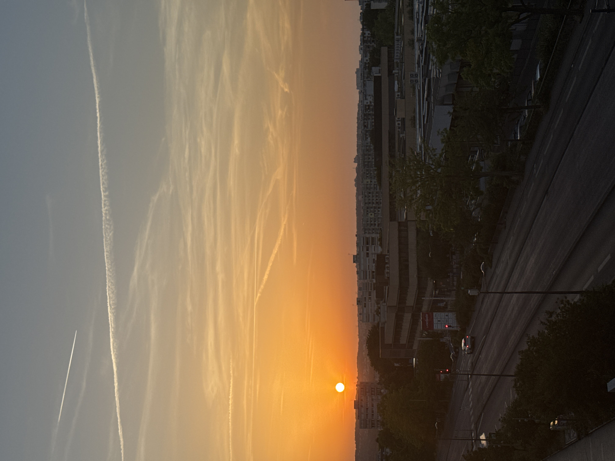 This is a vertical, eye-level, medium shot of a city at sunset. The sun, a bright orange disc, is low on the horizon, casting a warm glow across the sky. The sky is filled with wispy clouds and two distinct contrails from airplanes. In the foreground, a multi-lane road runs diagonally from the bottom left to the upper right of the frame. The road is flanked by trees and buildings. A single car is driving on the road, its brake lights illuminated. Traffic lights are visible, one red and one green. The buildings in the background are low-rise, with a mix of residential and commercial structures. The overall impression is one of a peaceful urban evening.