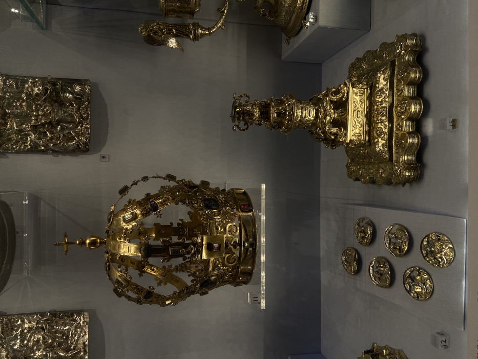 This image depicts an interior view of an exhibit at the Victoria and Albert Museum (V&A) in South Kensington, London, United Kingdom. The scene showcases an array of ornate, golden and silver-gilt artifacts displayed within glass cases and on plinths against a plain light gray wall.
Prominently featured in the upper-left portion is a large, elaborate golden crown. This open-work crown is adorned with numerous dark blue and red gemstones set within its arches and circlet, and is topped with a cross above an orb. It rests on a transparent acrylic stand, with a white numerical label "11" visible below it.
To the upper-right of the crown, a rectangular silver-gilt plaque with intricate figural relief work is mounted on the wall, identified by a white label "9". Further to the right, another similar relief panel is partially visible.
In the lower-right area of the image, a substantial and highly ornate golden candlestick or monstrance stands on a multi-tiered square base featuring detailed relief scrolls and foliage. This artifact is marked with a white label "15". Partially visible on the far right are additional golden objects, including one with a winged angelic figure and a lidded vessel, displayed on a white plinth.
In the lower-left, six oval-shaped golden medallions, each depicting a distinct figural scene in low relief, are arranged on a gray fabric pad within a glass display case, bearing the white label "12". The environment is characterized by controlled lighting, highlighting the metallic sheen and intricate details of the displayed objects. No individuals are present in the scene.
