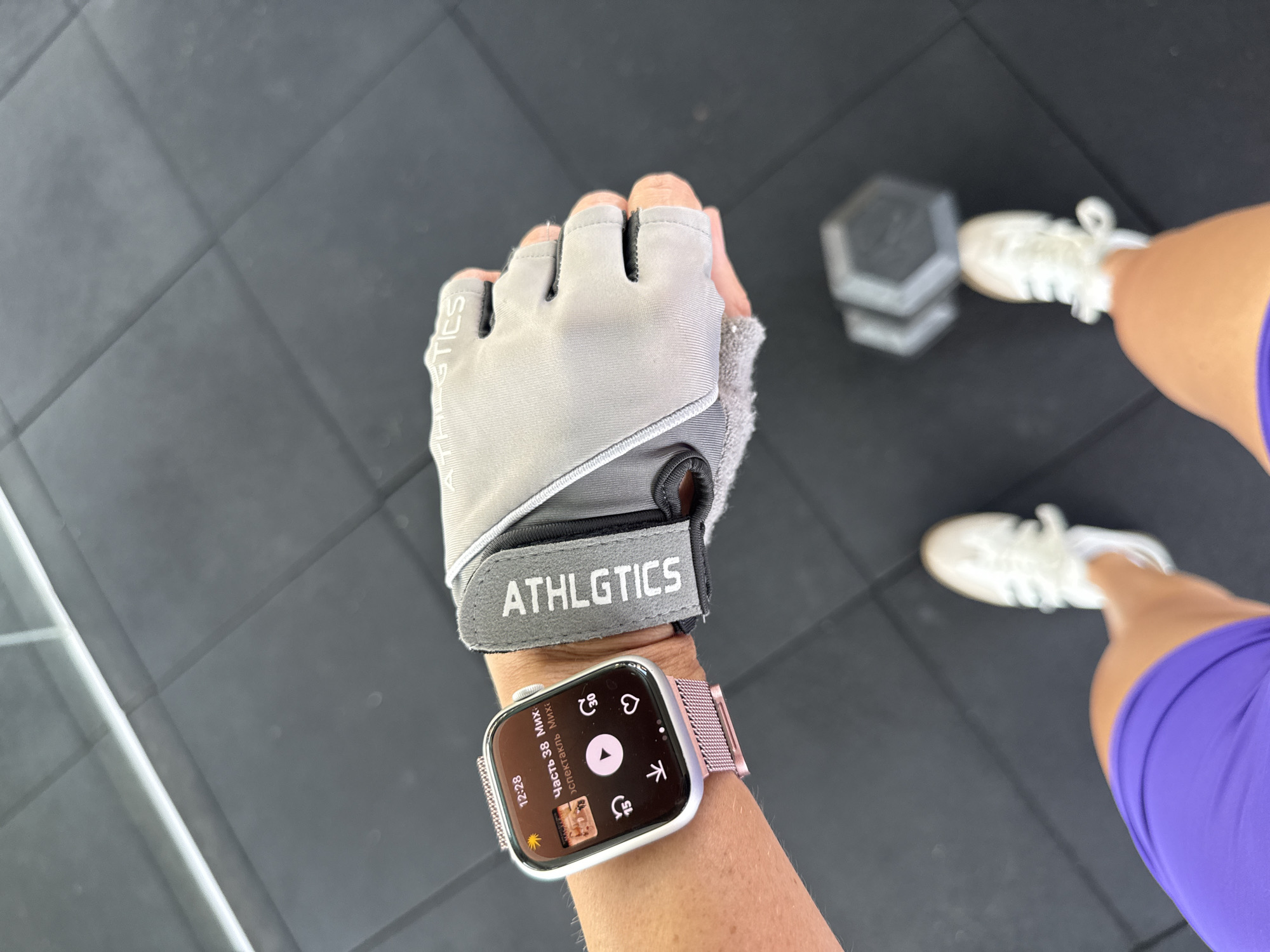 A first-person perspective shot shows a person's left arm wearing a silver Apple Watch with a pink Milanese loop band and a grey weightlifting glove with the word "ATHLGTICS" printed in white. The watch face displays the time as 12:28 and shows a music player interface with album art. The background is a black rubber gym floor with a visible hexagonal dumbbell to the right and a blurry view of white athletic shoes and purple shorts. The lighting appears to be natural, coming from above and to the left, suggesting an indoor gym setting. There is no indication of Kuta Selatan, Indonesia.