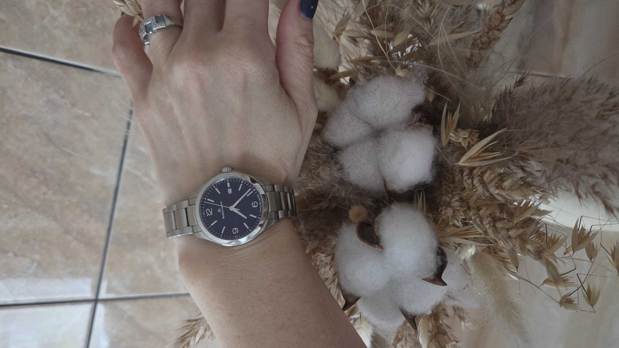 A close-up, overhead view captures a person's left arm and hand, adorned with a metallic watch and ring, resting beside a decorative dried floral arrangement. The hand features dark blue nail polish on the fingernails. On the wrist is a silver-toned Maurice Lacroix watch with a dark blue dial, displaying Arabic numerals at 12, 6, and 9, and a date window showing '20' at the 3 o'clock position. The brand name 'MAURICE LACROIX' is clearly visible on the watch face, along with minute markers including '05', '15', '30', and '45'. A silver-toned ring, set with square-cut light blue or green stones surrounded by smaller clear gems, is worn on a finger.
The dried arrangement is composed of fluffy white cotton bolls, various stalks of wheat, and different types of feathery dried grasses and seed heads in natural beige, brown, and off-white tones, creating a rustic aesthetic. The background consists of a light brown tiled floor with visible dark grout lines, suggesting an indoor setting, likely in a home in Chaikovsky, Russia. The lighting is bright and even, indicative of daytime. The image appears to be a posed shot, highlighting the accessories and the decorative elements.