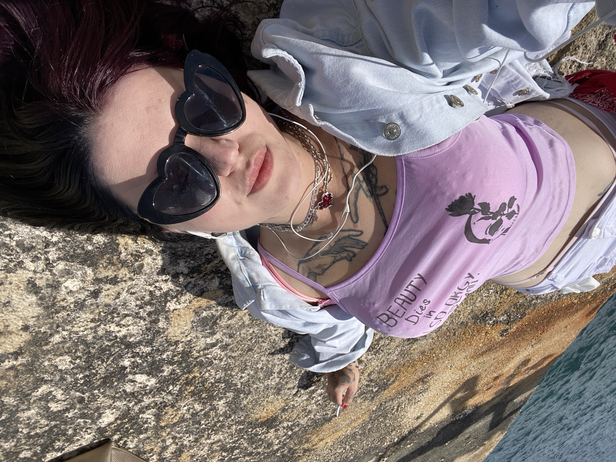 An overhead shot shows a woman lying on a rocky surface with water visible in the lower right corner. She has dark hair with reddish highlights, and is wearing heart-shaped sunglasses. She is dressed in a light purple tank top with the words "BEAUTY Dies in SD ORGY" printed on it. Over the tank top, she wears a light blue denim jacket. She also has several necklaces, a tattoo on her chest, and painted nails. Her right hand is visible and holds what appears to be a cigarette. The location is most likely the coastal area of the city Bar, Montenegro.