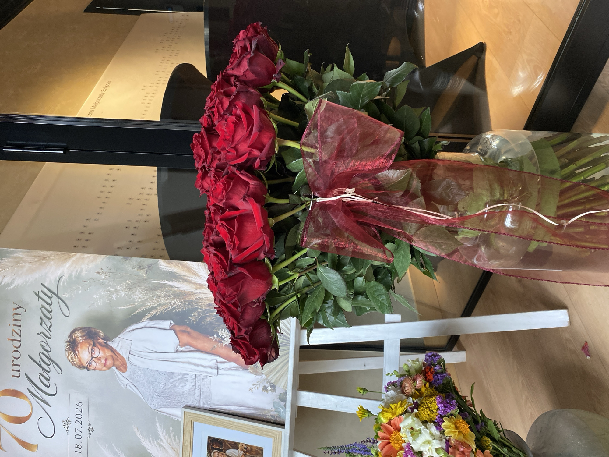 The image depicts a scene likely set indoors, possibly at a private residence or event space in Buk, Poland. In the foreground, a large bouquet of deep red roses is prominently displayed in a clear glass vase. The roses are tightly clustered and tied with a sheer, dark red ribbon. To the left, a smaller bouquet of mixed colorful flowers, including orange daisies, yellow flowers, and purple statice, is arranged in a grey vase.
Behind the flowers, a white easel supports a large portrait or banner. This banner features a photograph of an older woman with short blonde hair and glasses, wearing a light-colored outfit. Text on the banner indicates a 70th birthday celebration for "Małgorzata" and includes the date "18.07.2026." To the right of the banner, a portion of a dark, reflective surface, possibly a piano or a piece of furniture, is visible, reflecting the ambient light from the room. The background wall displays a repeating pattern of small, light-colored icons. The overall atmosphere suggests a celebratory occasion.