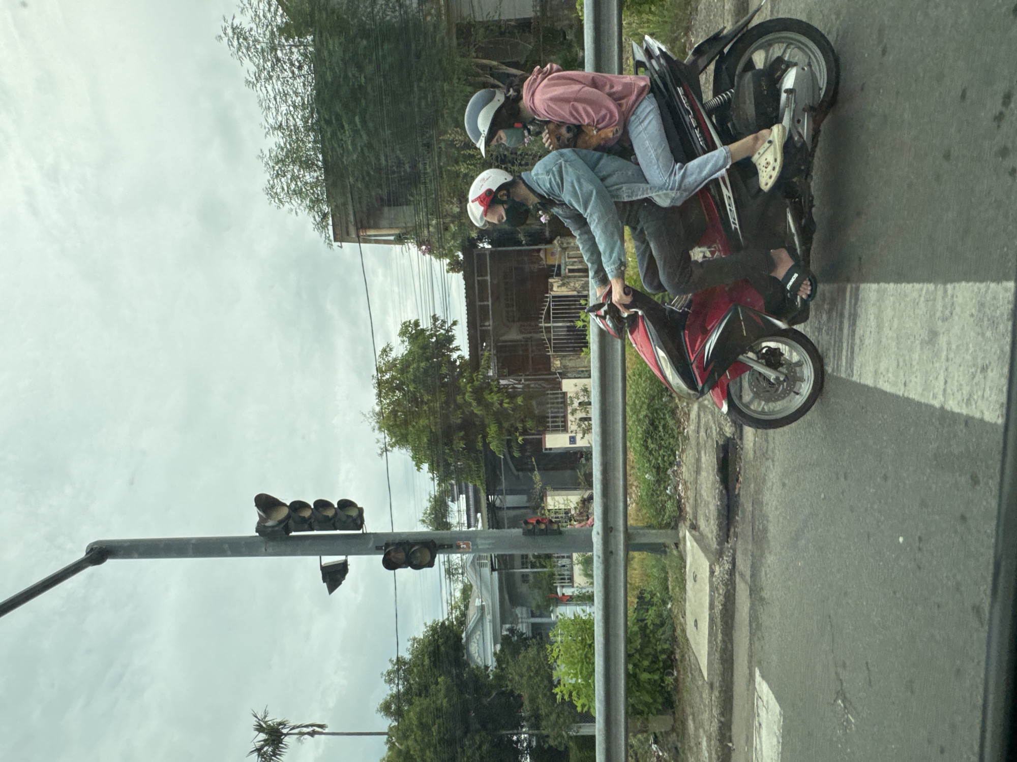 Two individuals are riding a red and black scooter on a street in Phú Xuân Ward, Vietnam. The driver, a male wearing a blue denim jacket, black pants, and a white helmet with a red stripe, has a black mask covering his face. The passenger, a female wearing a pink hoodie and blue jeans, has a blue helmet and a green mask. She is holding a small brown dog. Both individuals are wearing casual footwear. The scooter has "AIR BLADE 125" visible on its side. The street appears to be asphalt with white crosswalk markings. In the background, a traffic light pole with multiple signals is visible. Beyond the traffic light, there are houses, trees, and vegetation. The sky is overcast.
