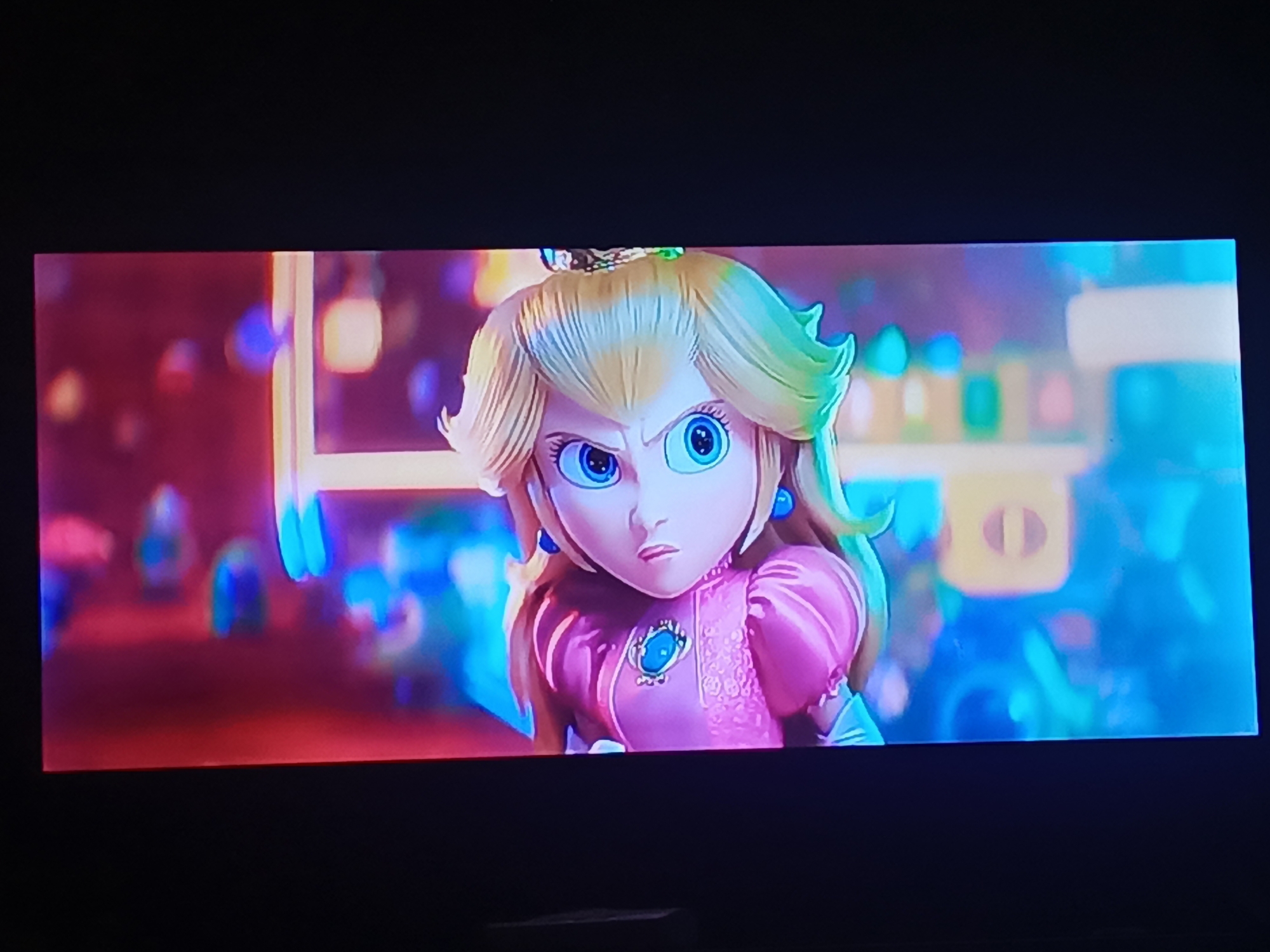 The image depicts an animated character, Princess Peach, from the Super Mario Bros. franchise. She is shown with a stern expression, furrowed brows, and wide eyes. Peach is wearing her signature pink dress with a blue brooch and blue earrings. Her blond hair is styled with curls. The background consists of blurred lights and shapes suggesting an urban or amusement park setting. There is no indication of the location being Juan Griego, Venezuela, within the image.